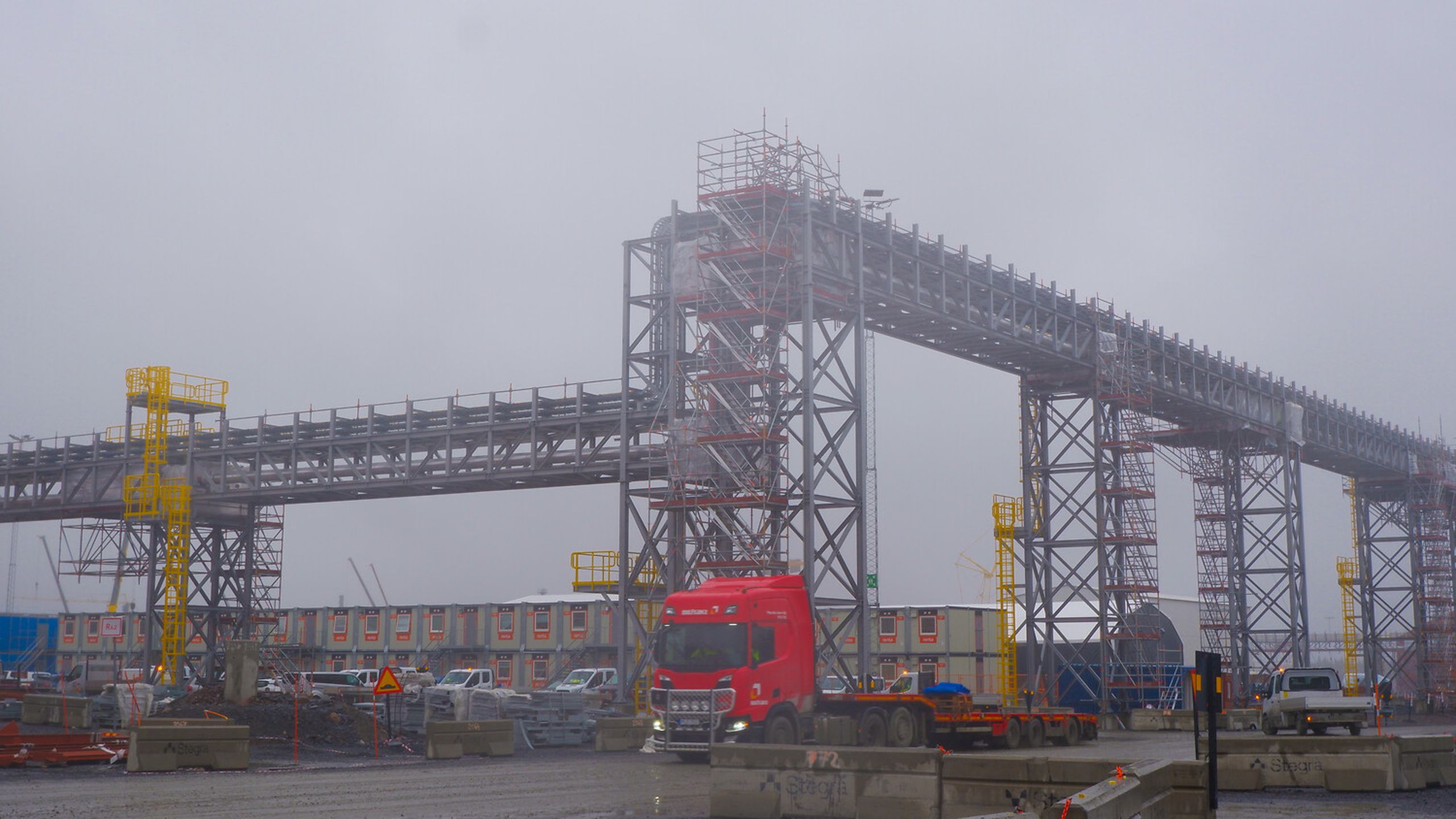 A red truck drives under large metal structures at an industrial site on a cloudy day, with construction equipment and workers visible.
