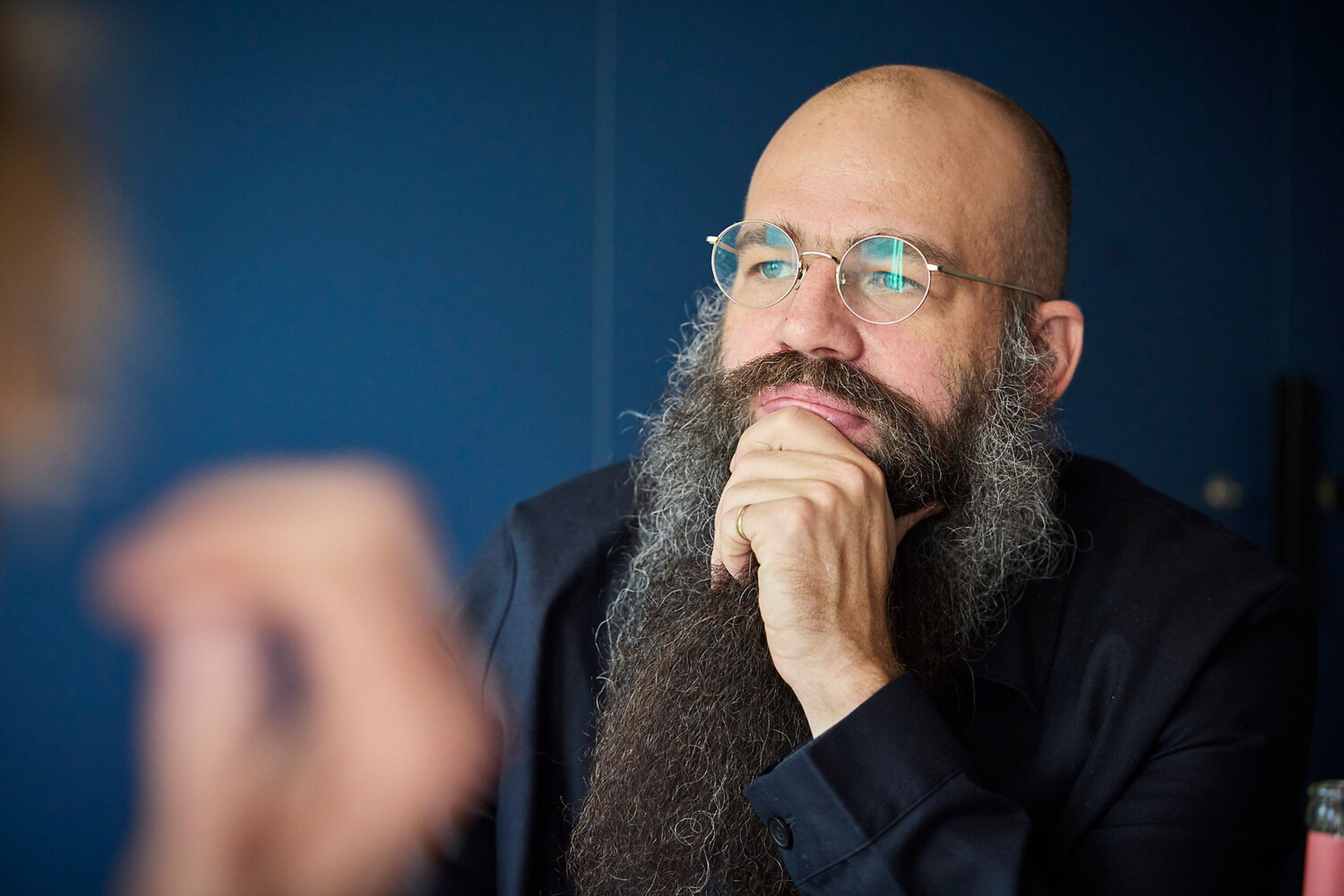 A man with a long beard and glasses, wearing a dark shirt, rests his chin on his hand, looking thoughtfully to the side.