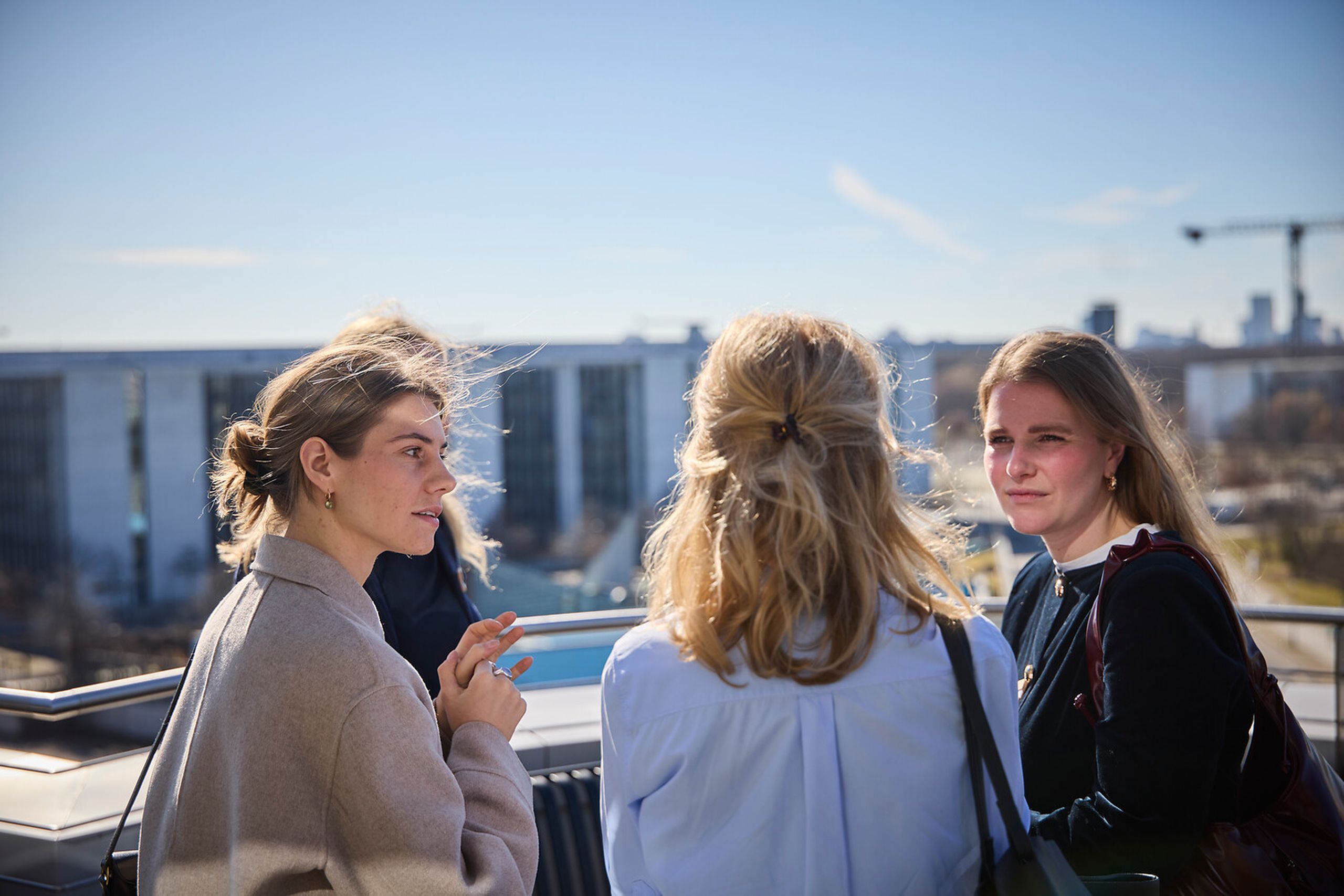 Three women talking outdoors on a sunny day with a cityscape and construction crane in the background.