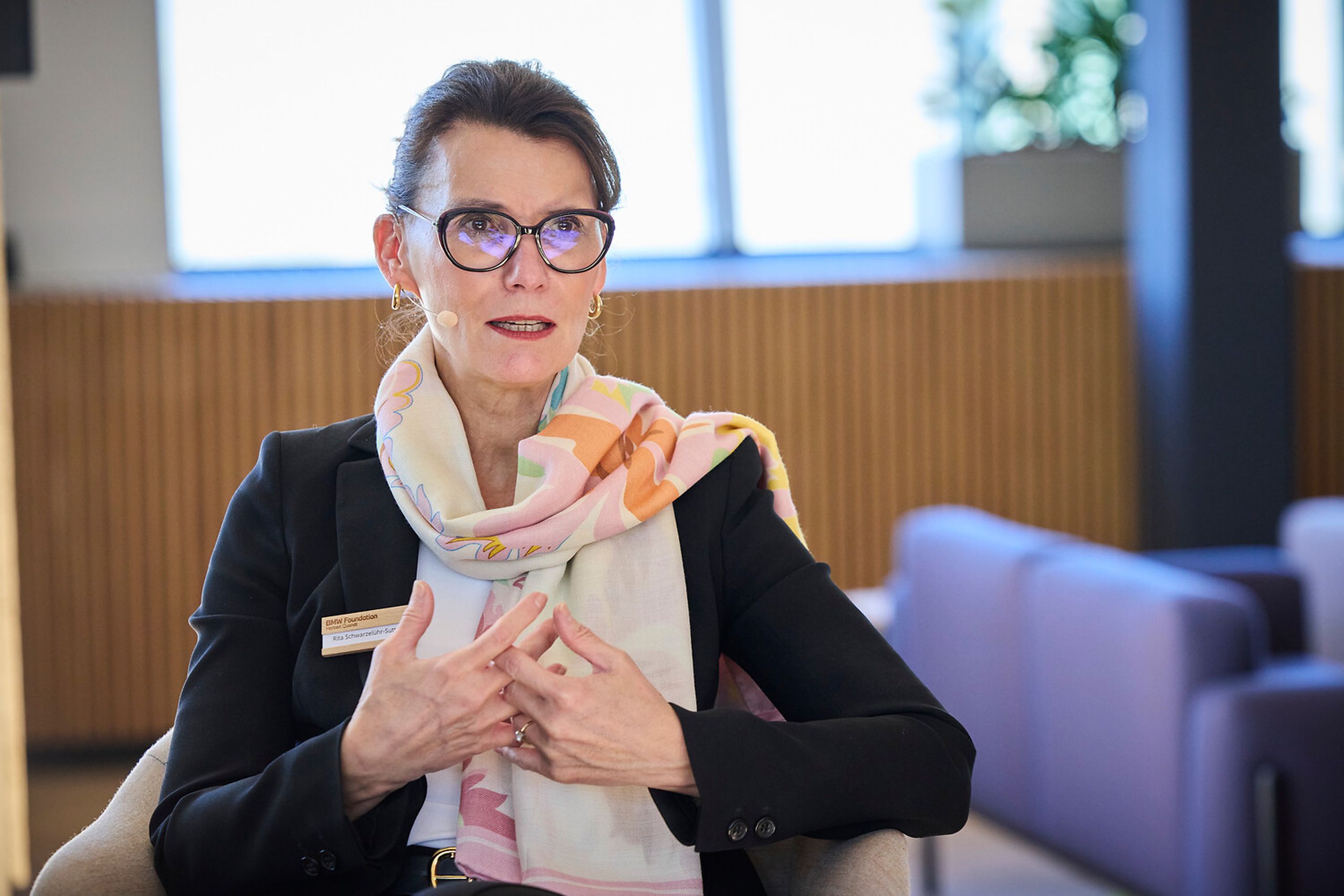 Woman with glasses and floral scarf speaks, gesturing with hands in a modern office setting, with large windows and upholstered chairs.