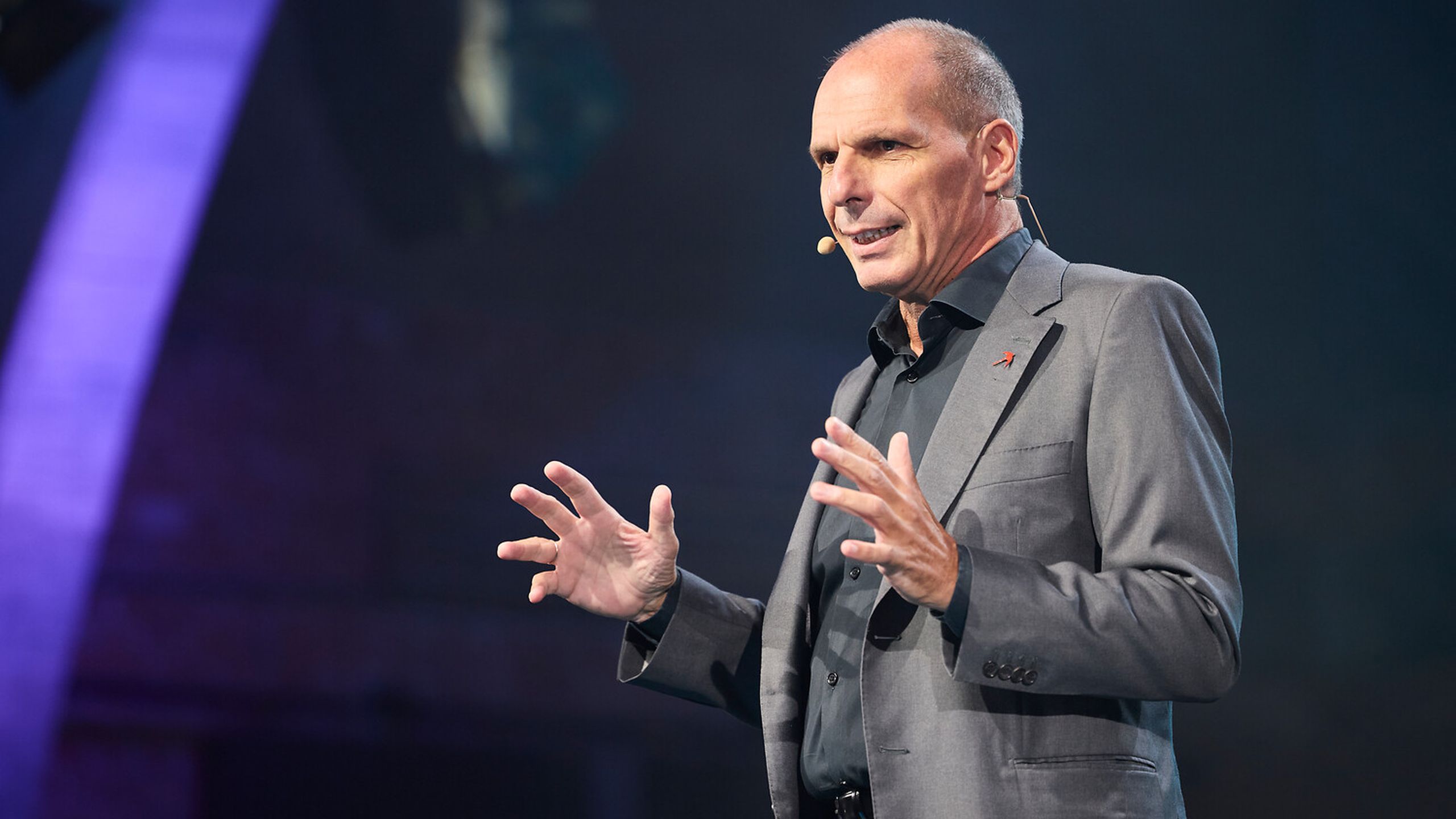 A person in a gray suit speaks passionately on stage with a headset microphone, gesturing with both hands.