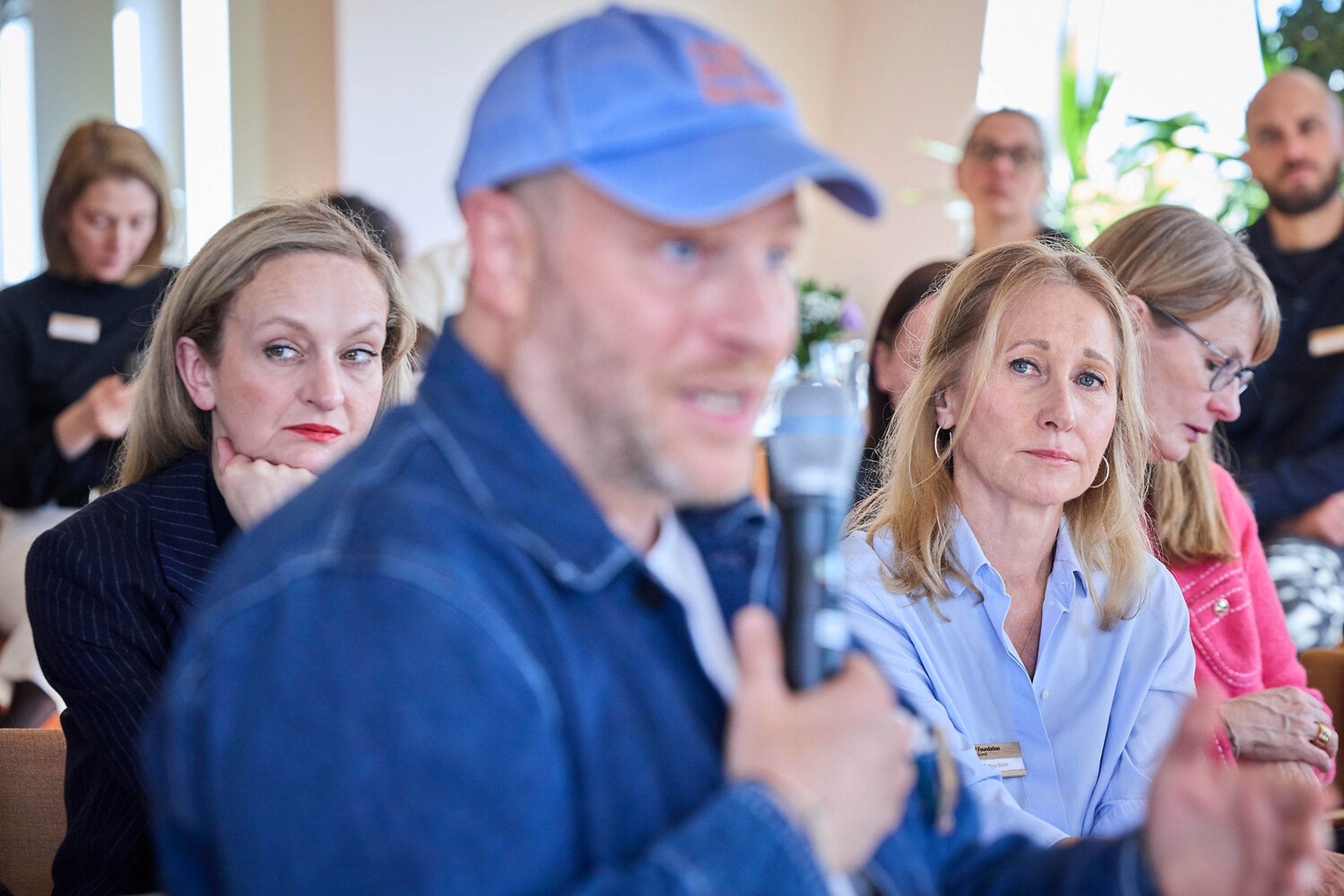 A man in a blue cap speaks into a microphone in a meeting, while two women focus intently on him. Other attendees are visible in the background.