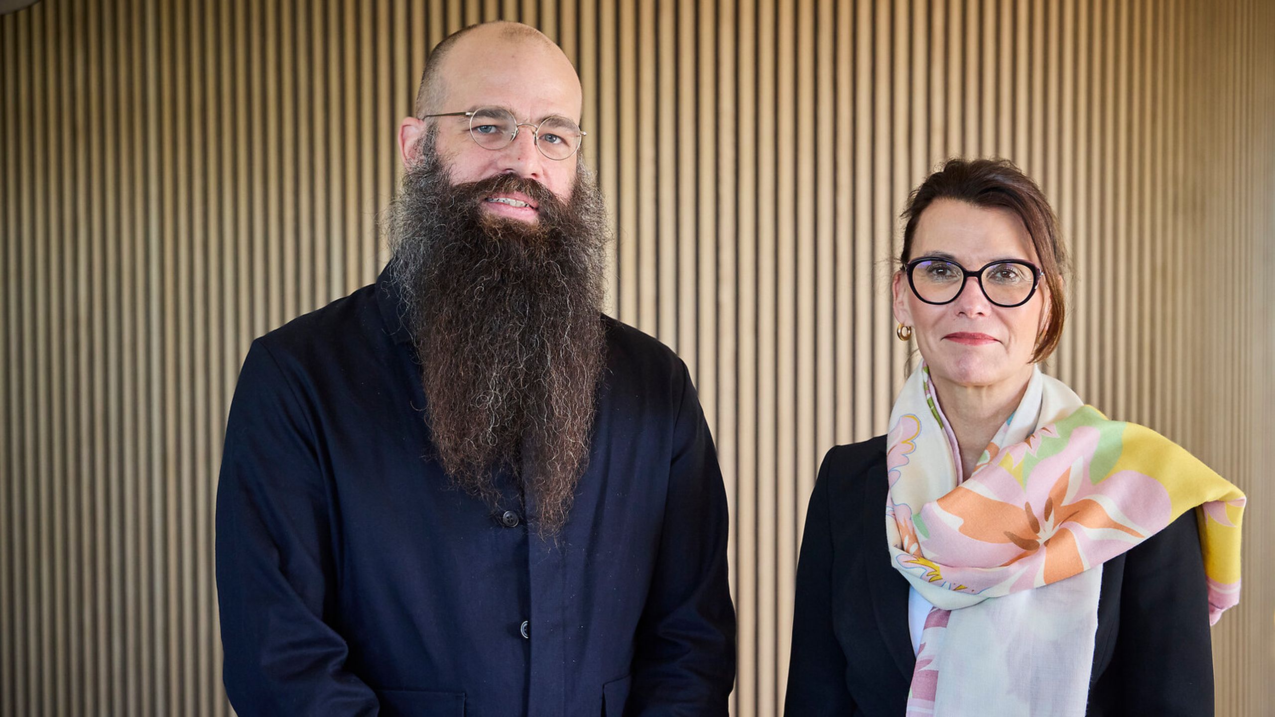 Two people in formal attire stand in front of a wooden panel wall. One has a long beard, and the other wears glasses and a colorful scarf.