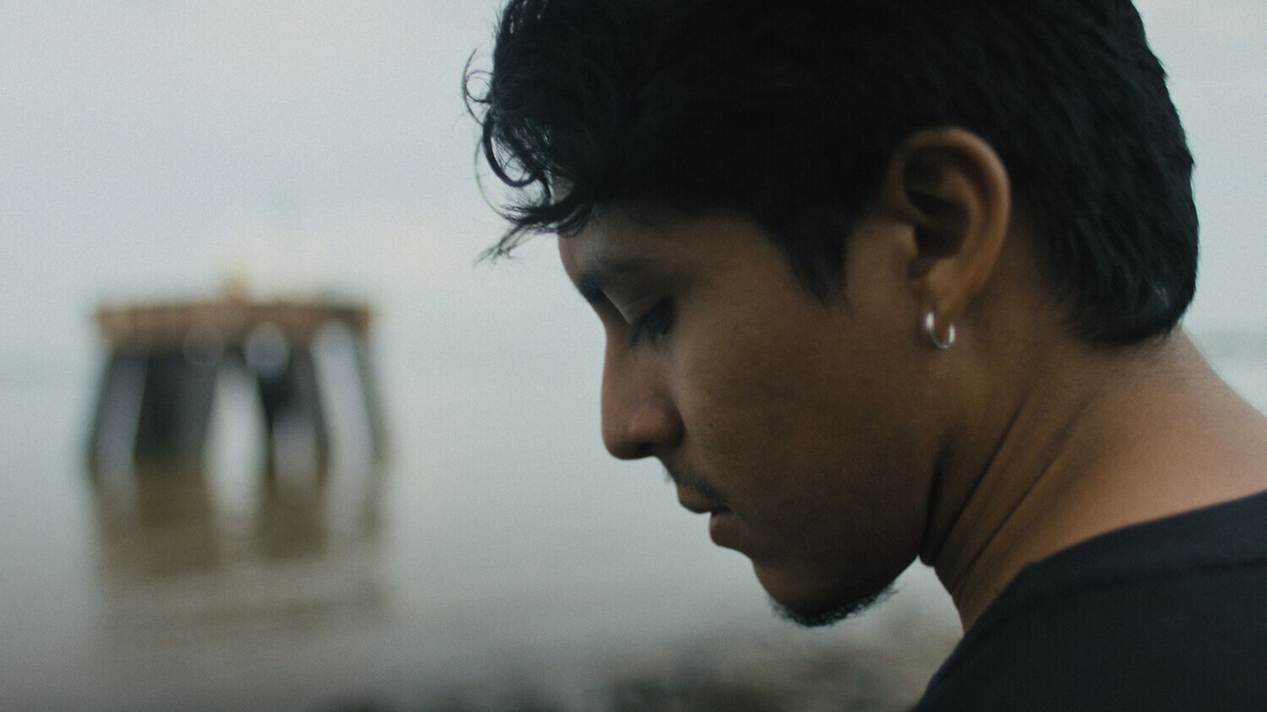 A person with short hair and an earring looks down, standing near a calm body of water with a distant pier in the background.