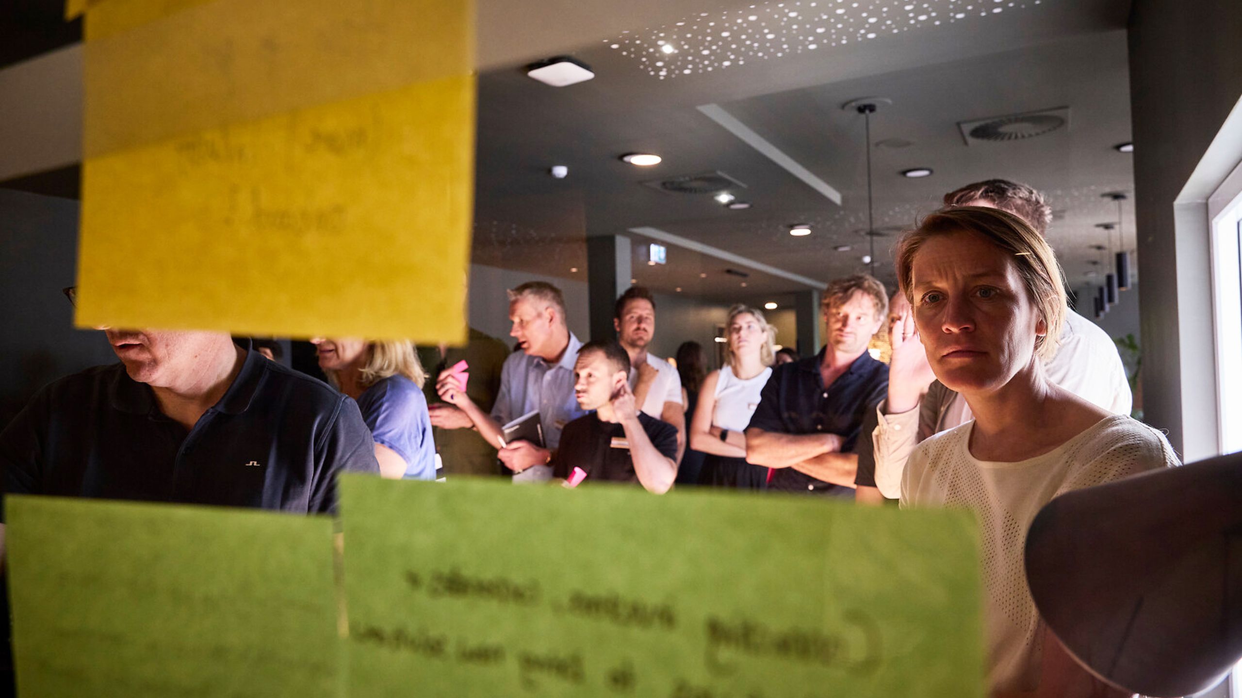 A group of people in a room, focused on colorful sticky notes on a glass wall, participating in a brainstorming session. A group of people in a room, focused on colorful sticky notes on a glass wall, participating in a brainstorming session.