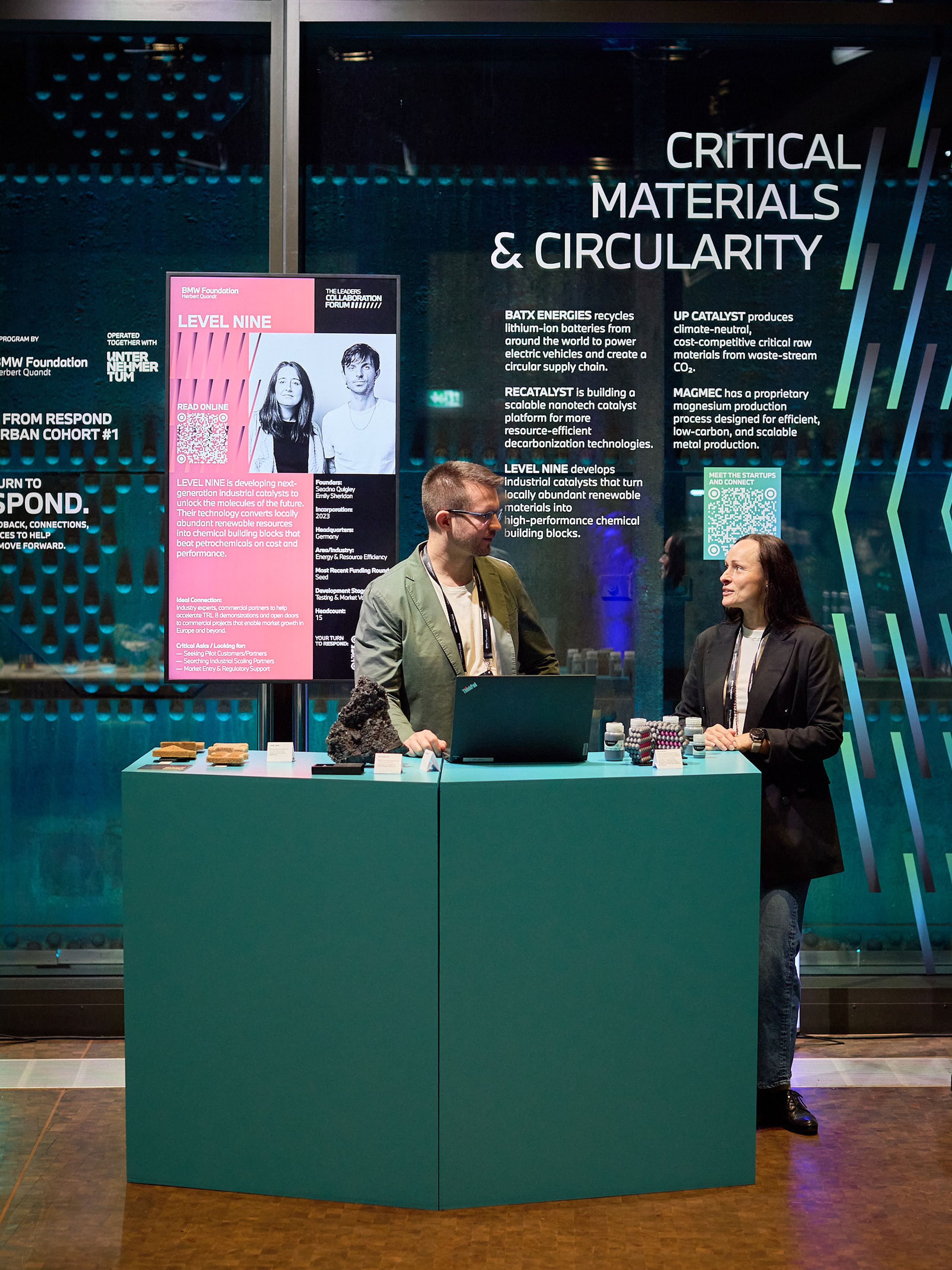 Startups from the BMW Foundation RESPOND accelerator showcased their cleantech solutions at the Forum. Two people stand behind a blue desk with a laptop and brochures, in front of a display about critical materials and circularity.