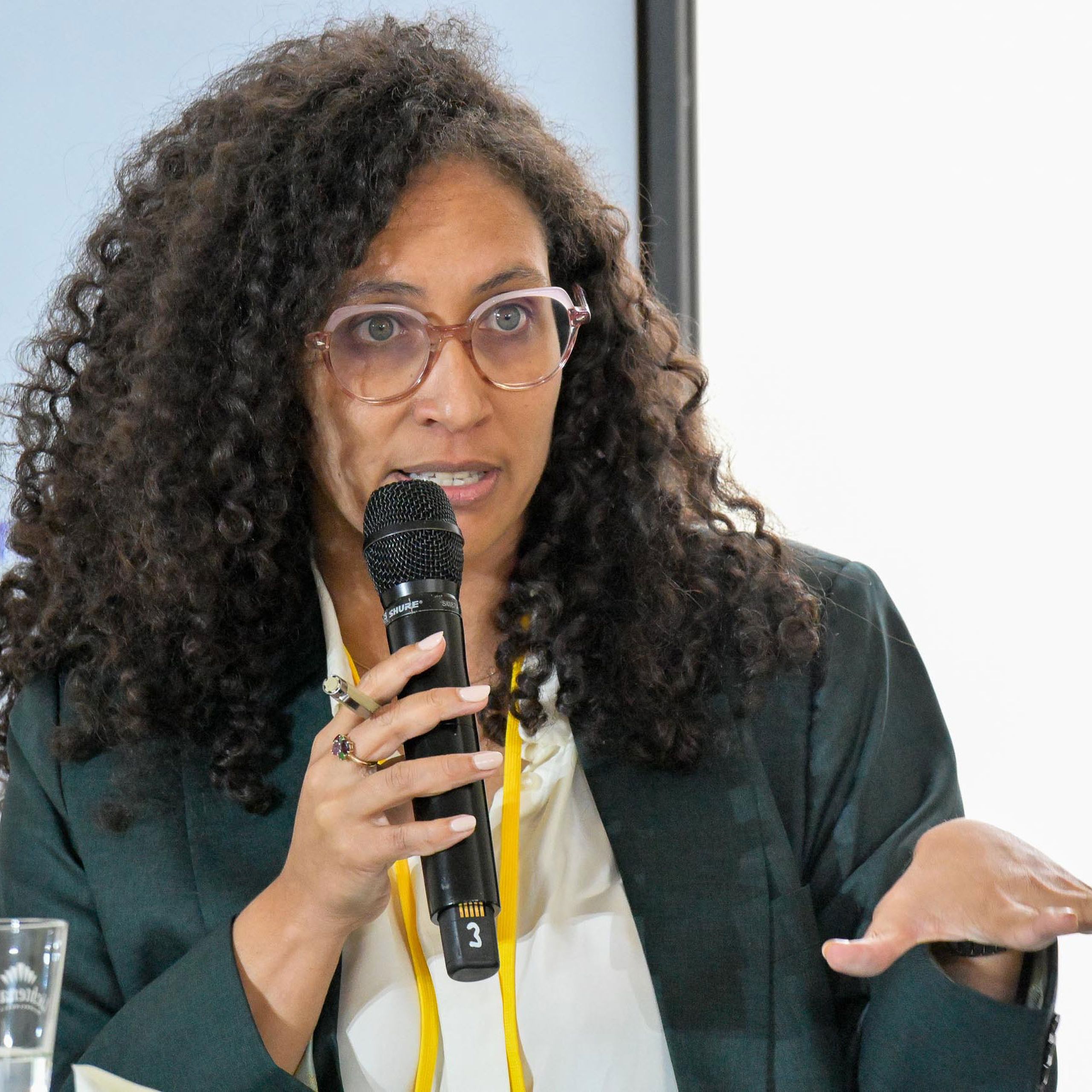 A woman with curly hair and glasses speaks into a microphone, gesturing with her hand, during a panel discussion.