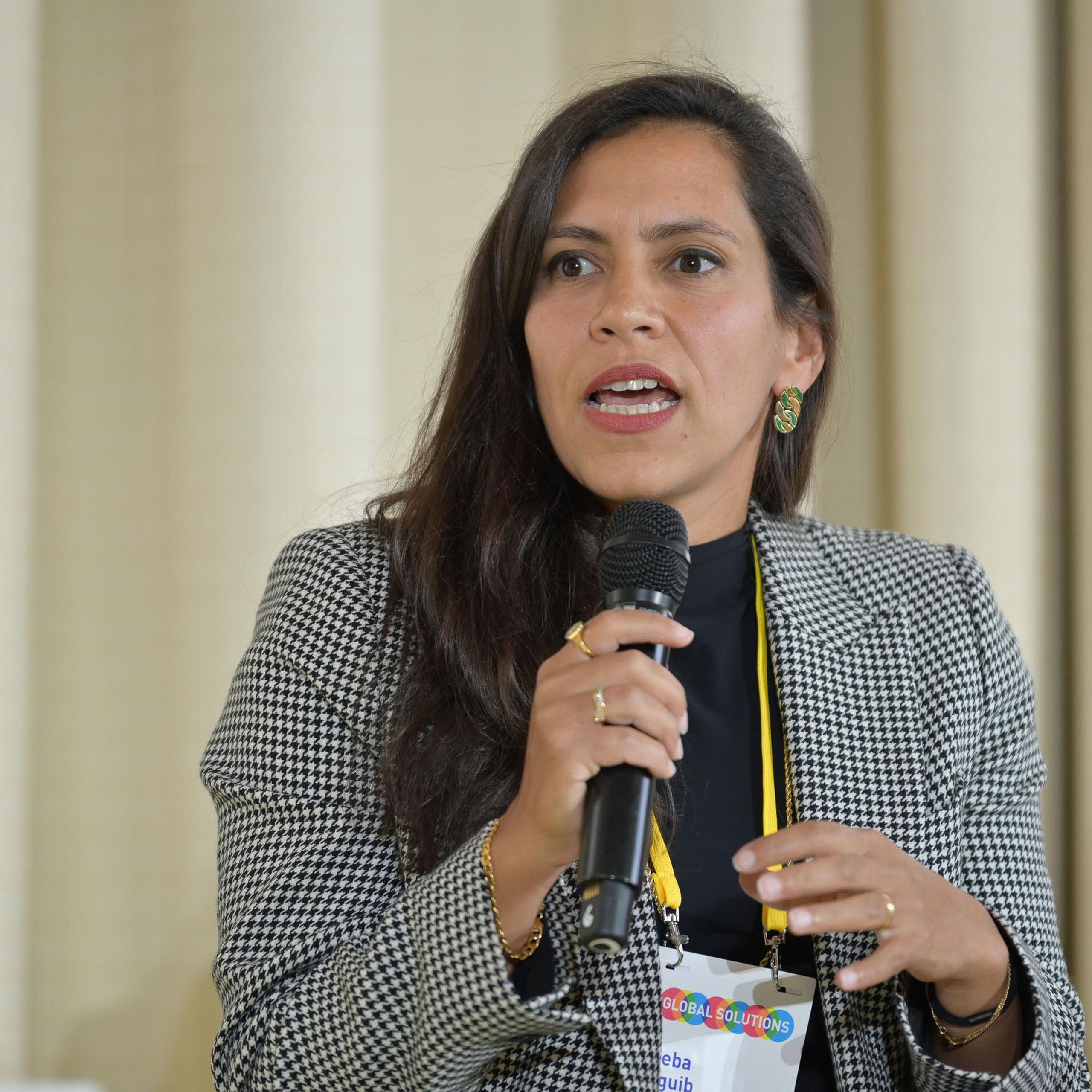 Woman speaking into a microphone, wearing a houndstooth blazer and conference badge, gesturing with her hand.