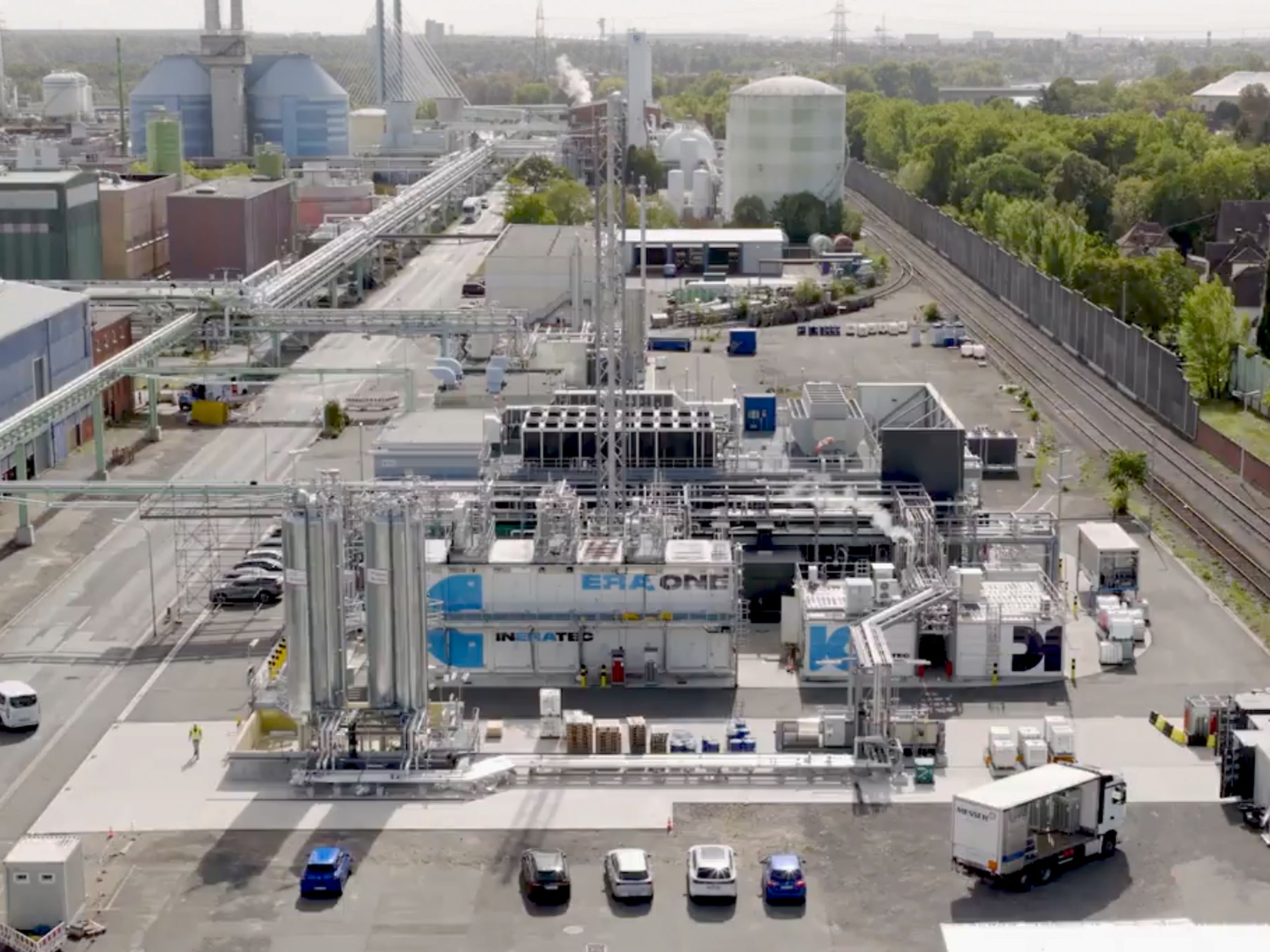 With the pioneer plant ERA ONE in Frankfurt, Ineratec is ramping up operation of the largest Power-to-Liquid plant to date for the production of synthetic, climate-friendly e-Fuels An industrial complex with pipes, buildings, and vehicles, including trucks and cars, located next to a railway line and surrounded by trees.