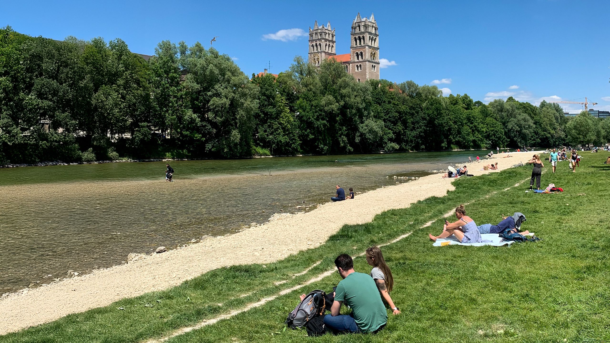People relaxing on a grassy riverside with a sandy bank, trees, and a large building with towers in the background under a blue sky. People relaxing on a grassy riverside with a sandy bank, trees, and a large building with towers in the background under a blue sky.