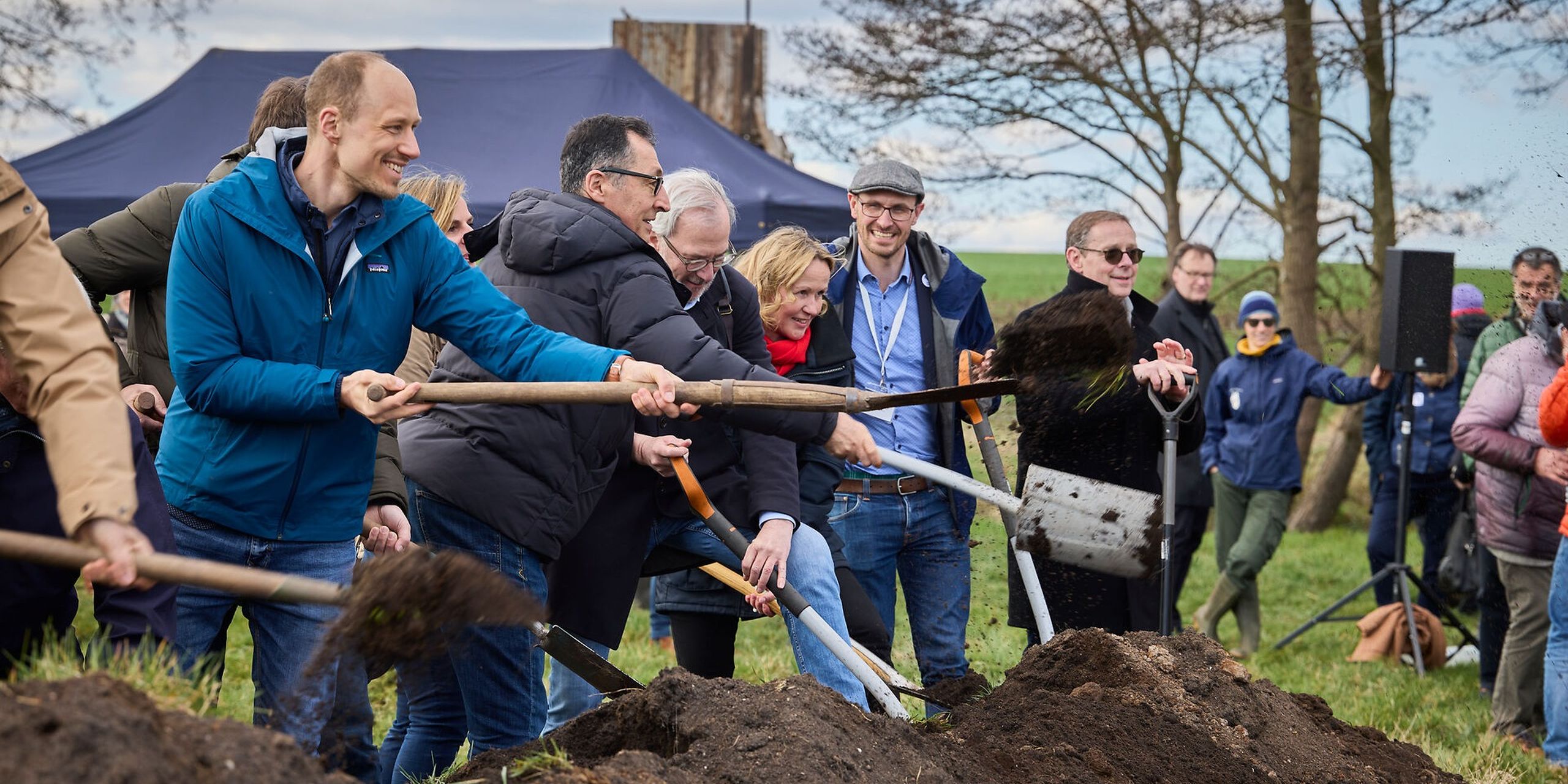 A group of people smiling and using shovels to dig soil at an outdoor event, with a tent and trees in the background.