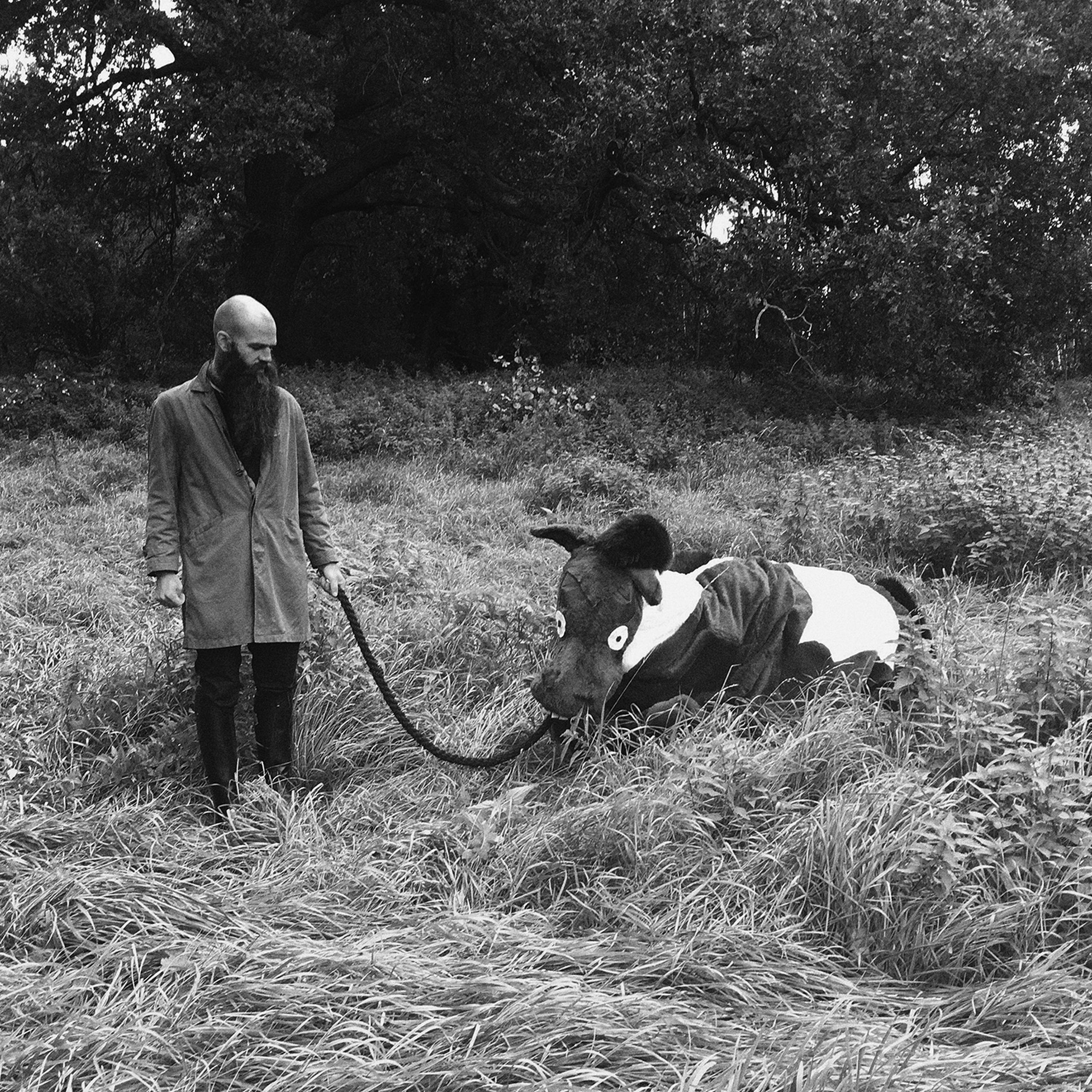 A person in a coat leads a tapir on a leash through a grassy field with trees in the background, in a black and white photo.