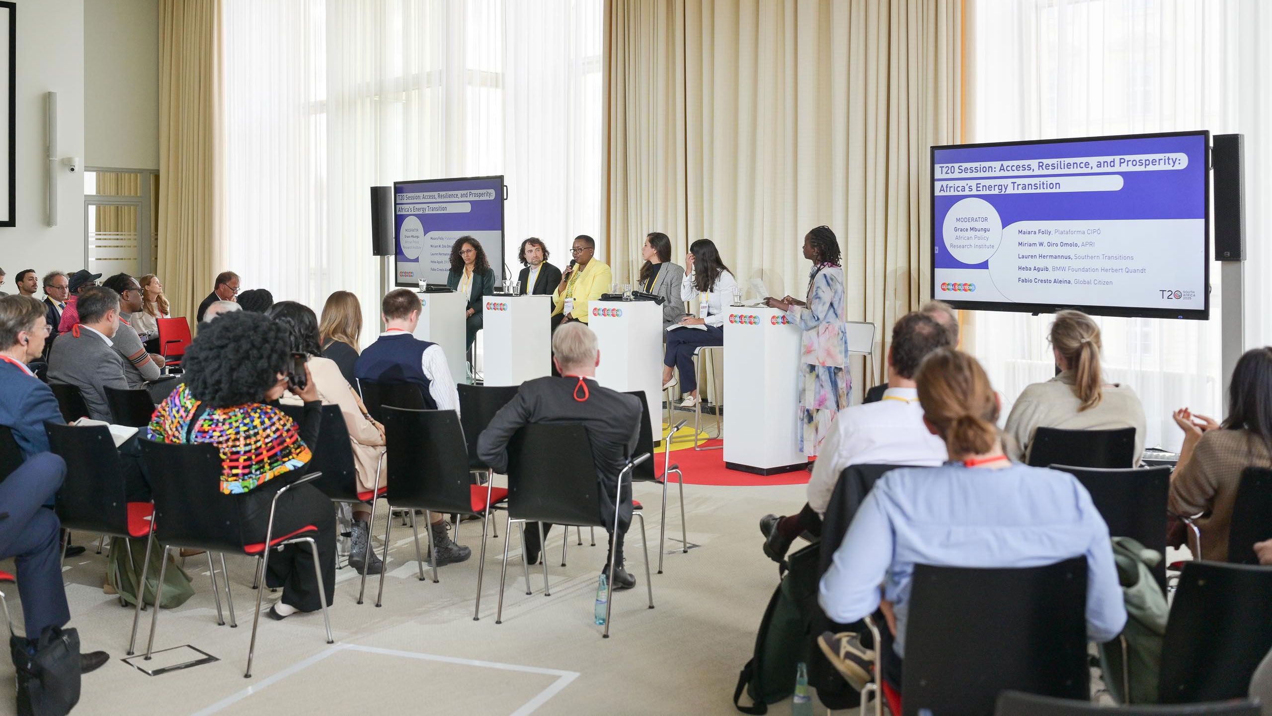A panel of seven speakers discusses energy transition at a conference. Attendees sit facing them, and a presentation screen is visible.