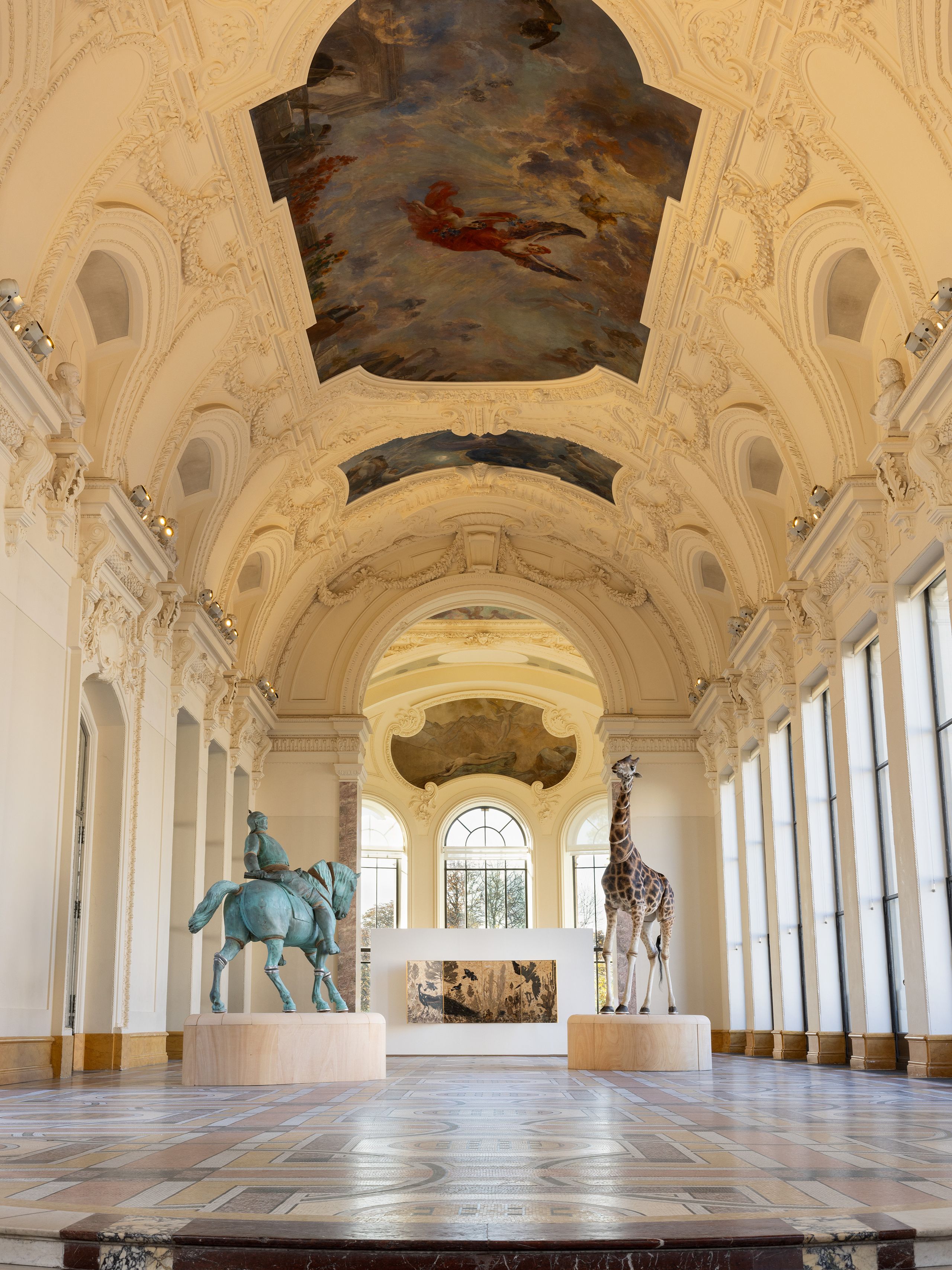 Opulent gallery with ornate ceiling, statues of a horseman and a giraffe, large windows, and a detailed floor pattern.