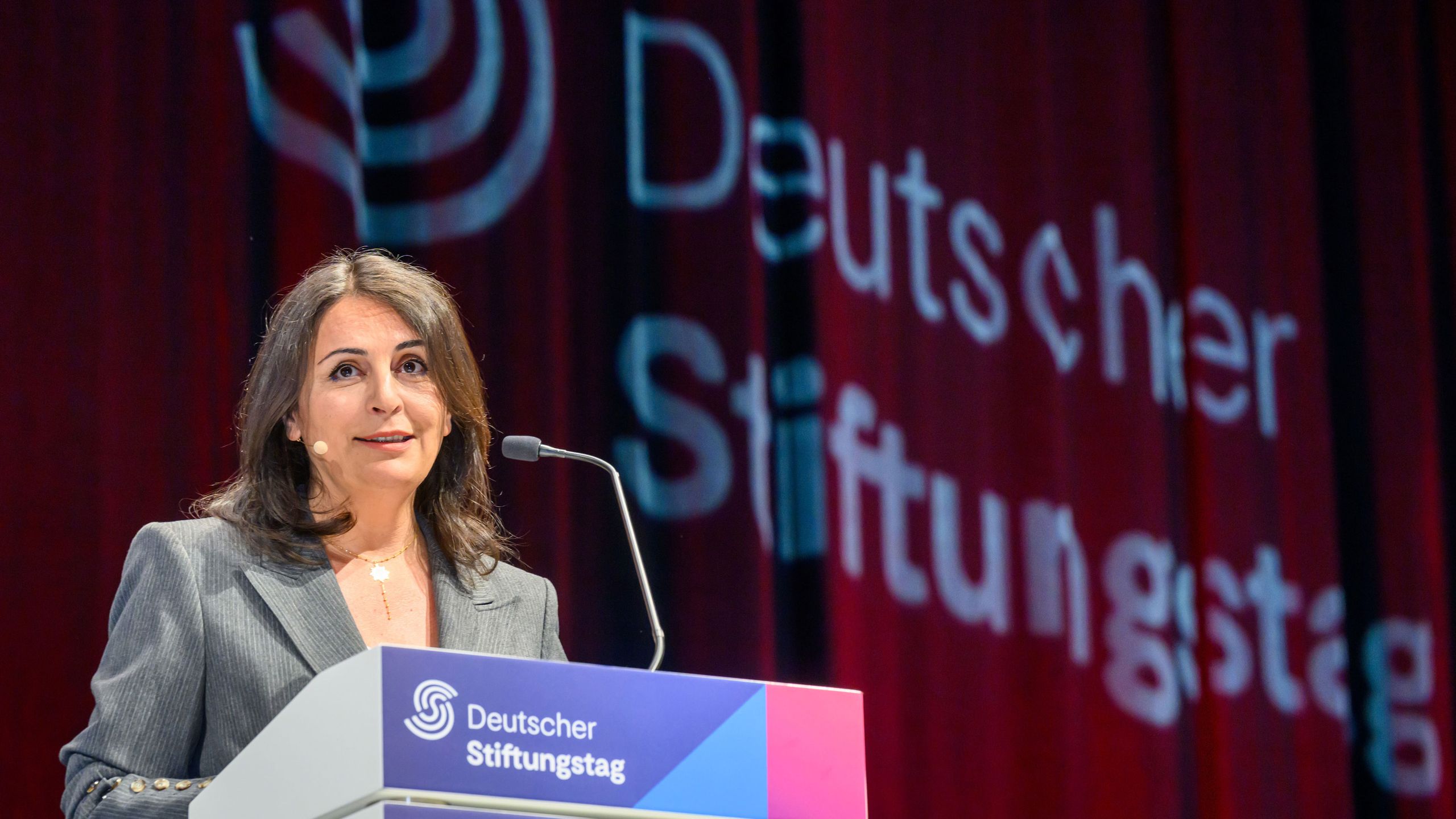 A woman speaks at a podium with "Deutscher Stiftungstag" projected on a red curtain in the background.