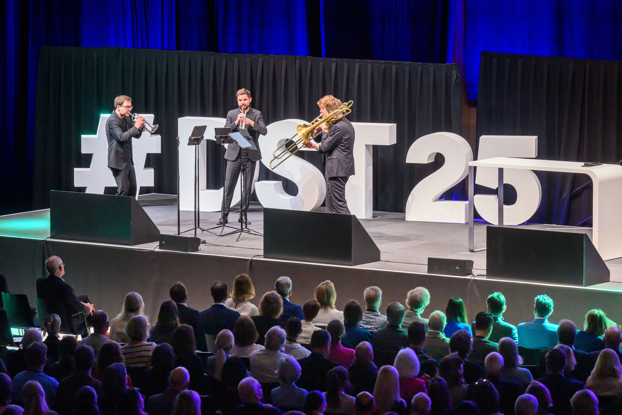 Three musicians play brass instruments on stage with large "#LDST25" letters in the background, while an audience watches.