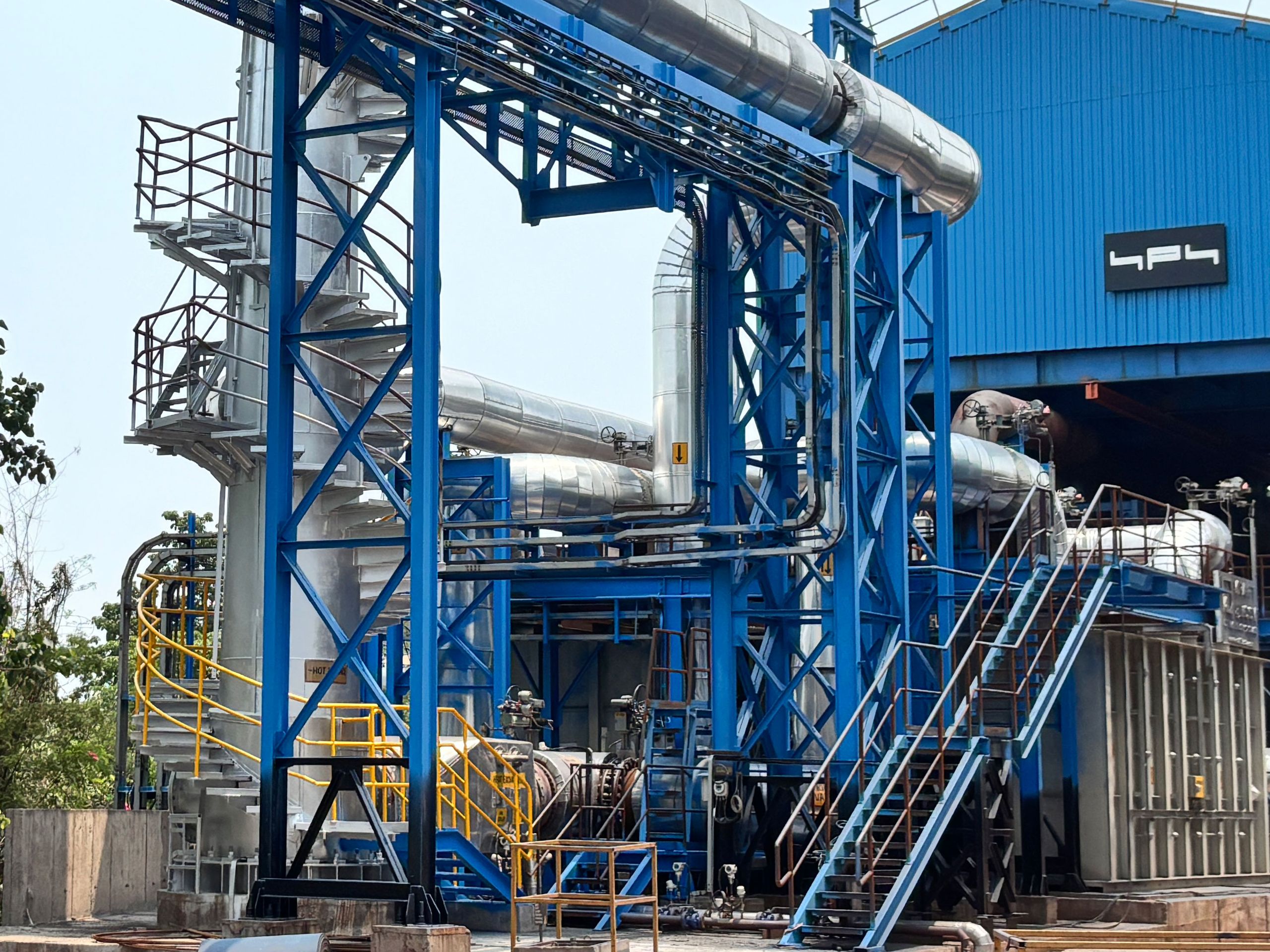 At one of the first steel mills from Tata Steel Limited in Jamshedpur, India, Kraftblock deployed a waste heat project recovering heat from the sinter cooler of Sinter Plant 3. Industrial facility with blue steel structure, interconnected pipes, and a building in the background, set in an outdoor environment.