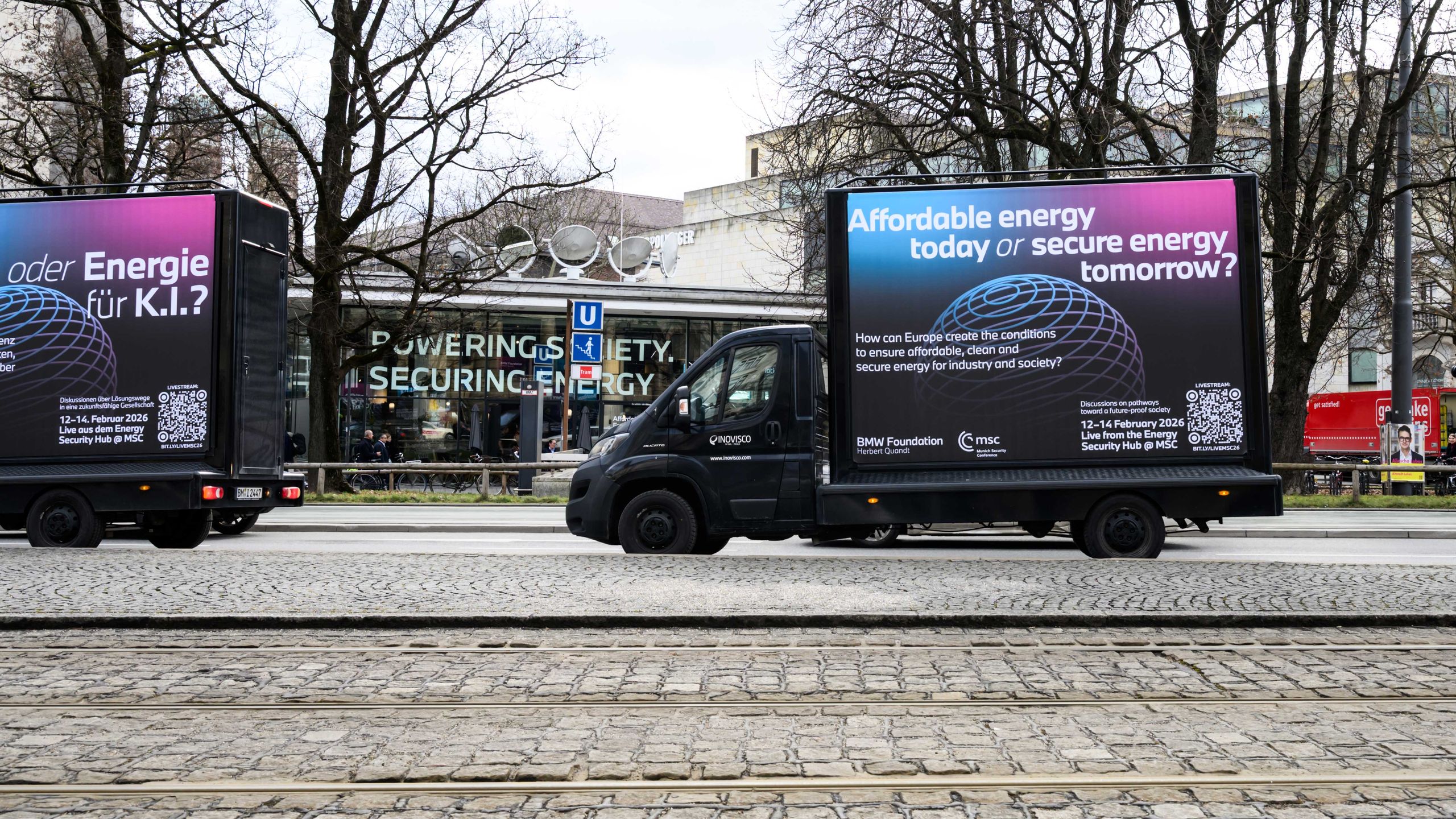 Two trucks display contrasting energy messages, parked on a cobblestone street with bare trees in the background.