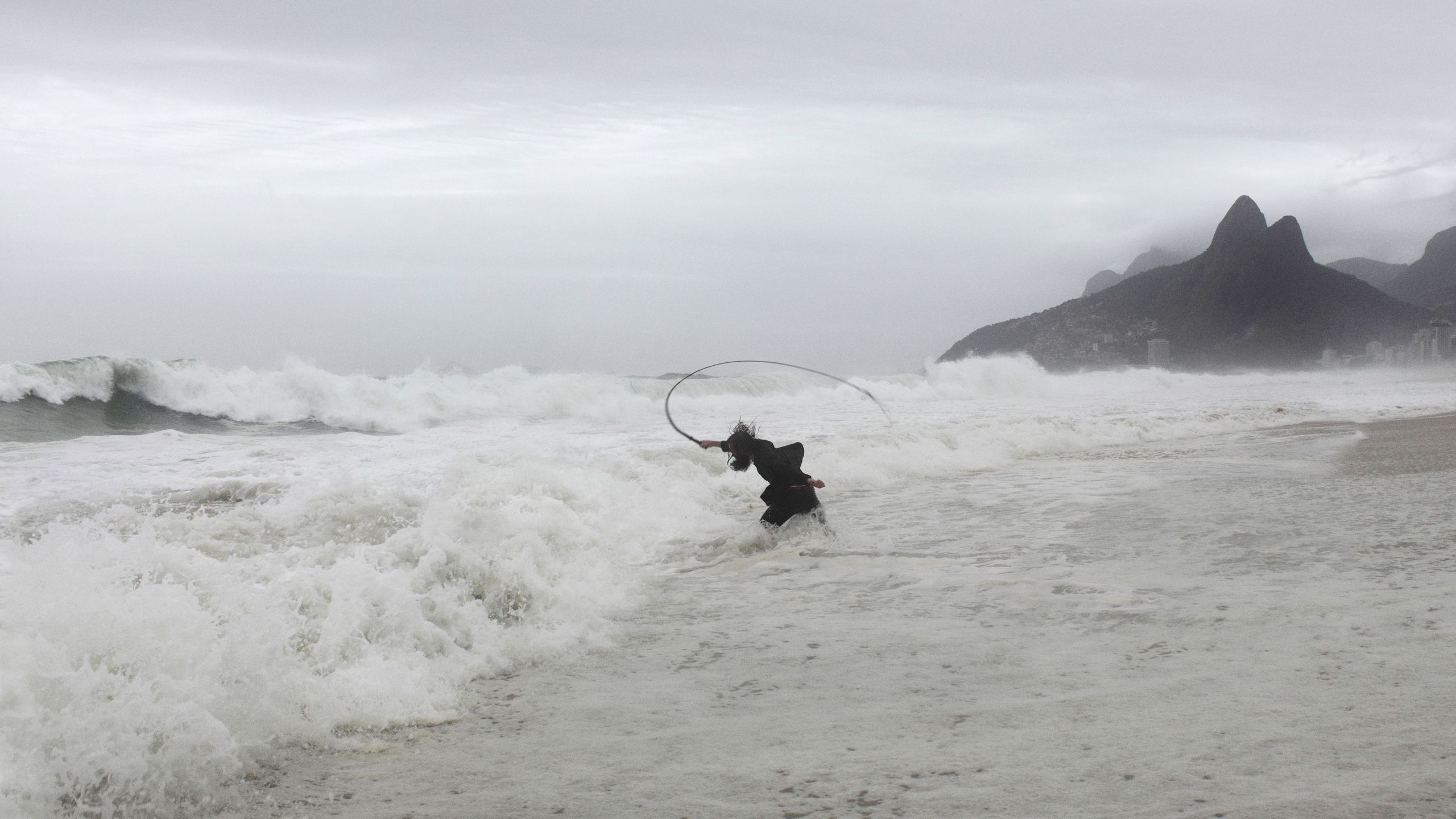 Person casting a fishing net in turbulent ocean waves near a rocky coastline; overcast sky and rough sea conditions.
