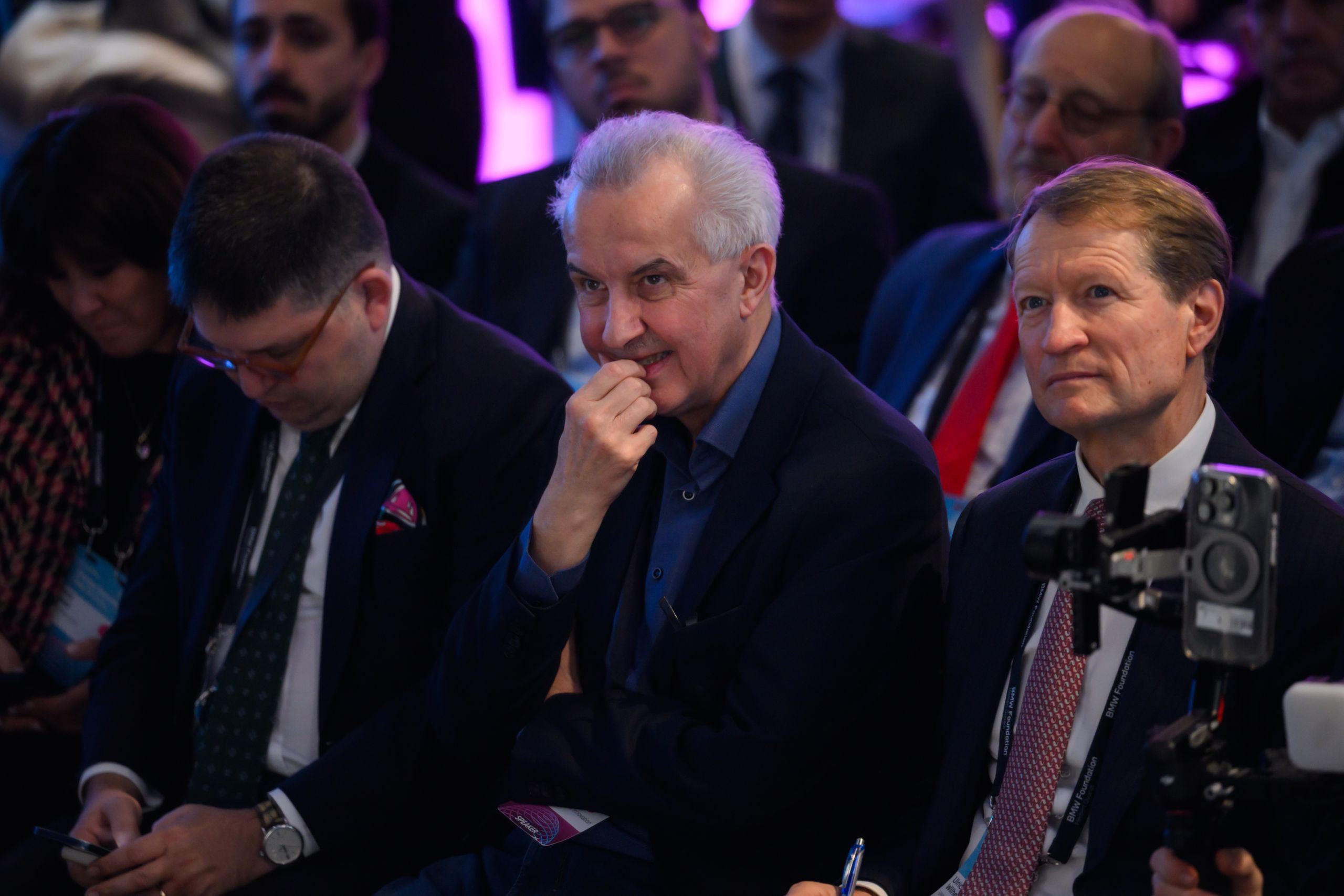 People seated in an audience at a conference, with focus on three men in suits in the front row, one holding his chin thoughtfully.