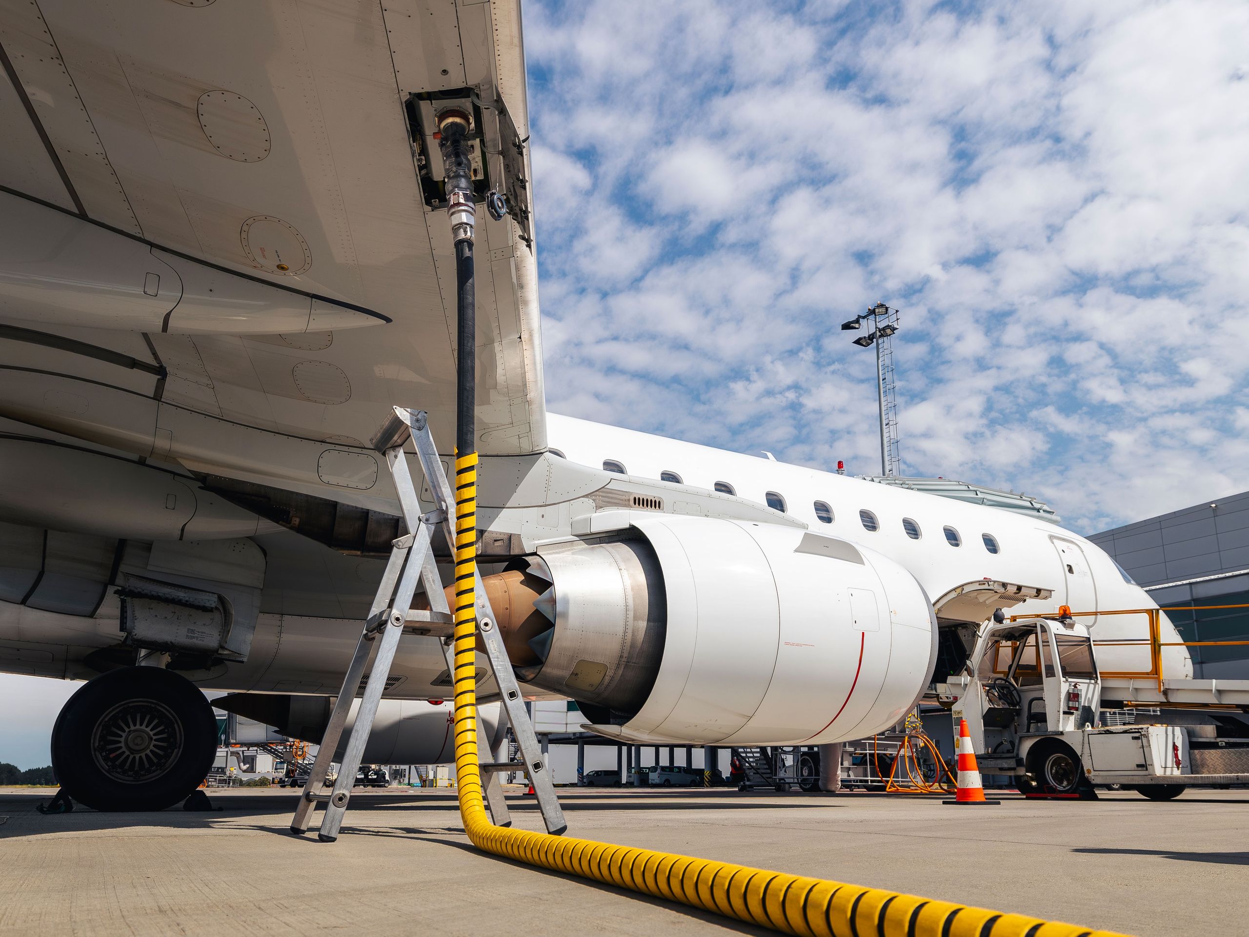 Airplane parked on a runway at an airport, receiving supplies. Burning just one kilogram of kerosene releases approximately 3.16 kilograms of CO₂. Airplane parked on a runway at an airport, receiving supplies via a yellow hose, with a clear sky and scattered clouds above.