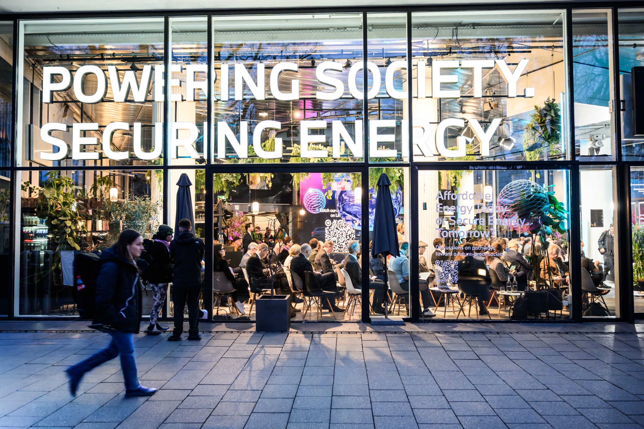 People sitting in a glass-walled building with large signs reading "Powering Society" and "Securing Energy" above them.