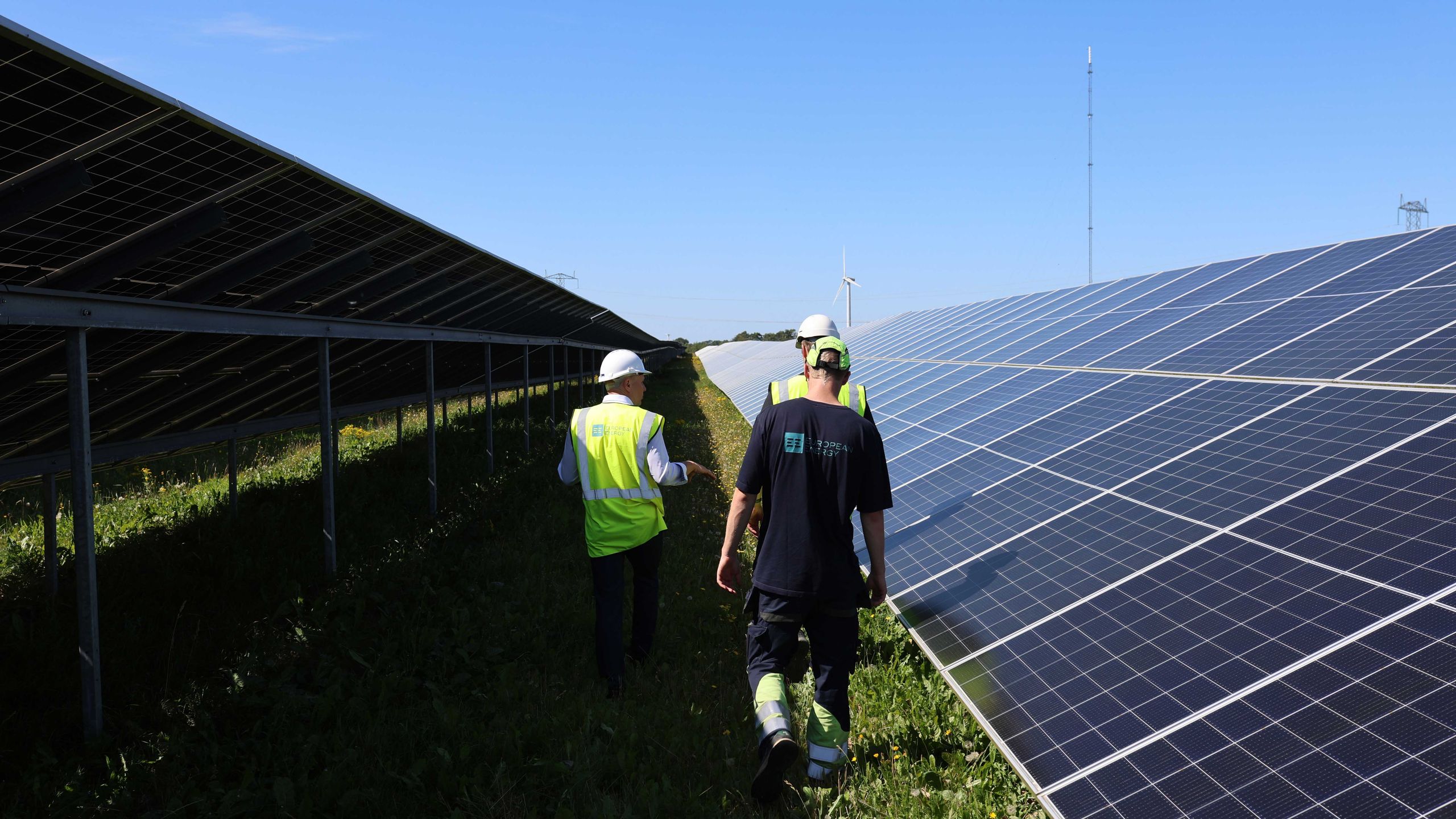 Three people in work attire walk between rows of large solar panels under a clear blue sky. Three people in work attire walk between rows of large solar panels under a clear blue sky.