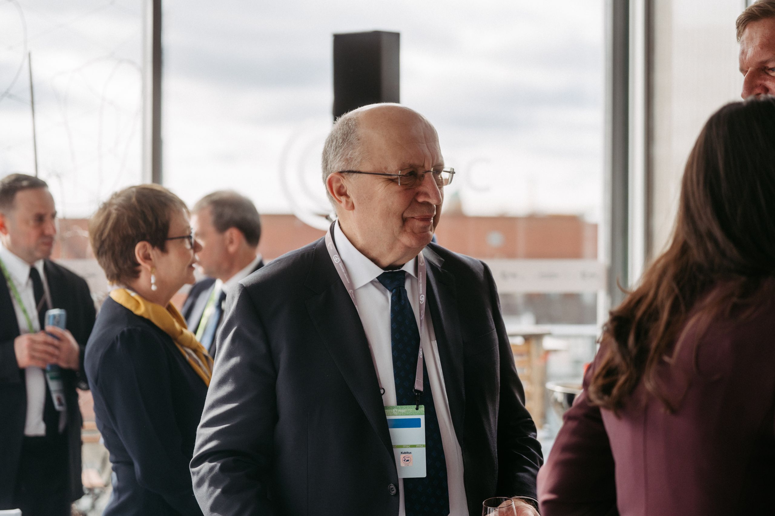 A group of people in business attire engage in conversation at an indoor event with large windows in the background.