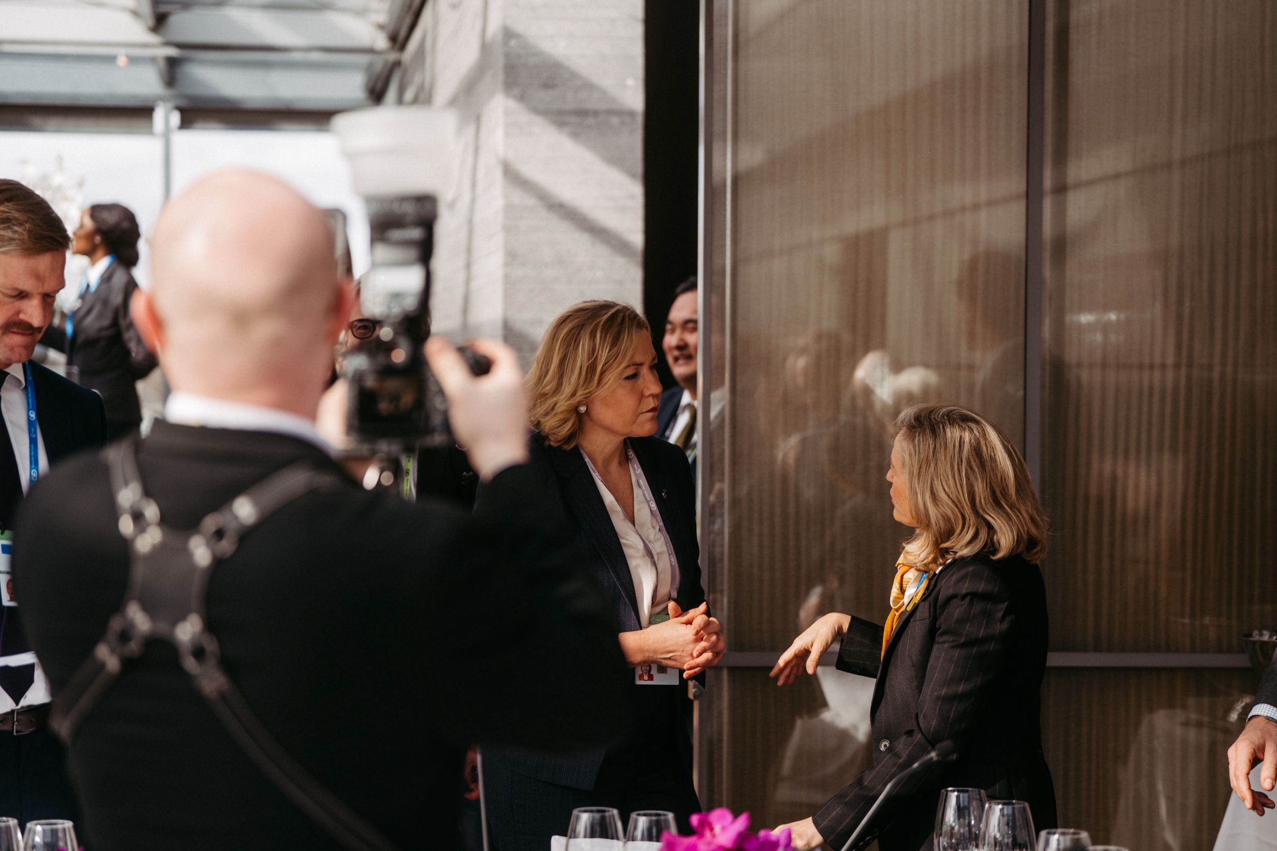 People in formal attire are having a discussion indoors, with a photographer capturing the moment.