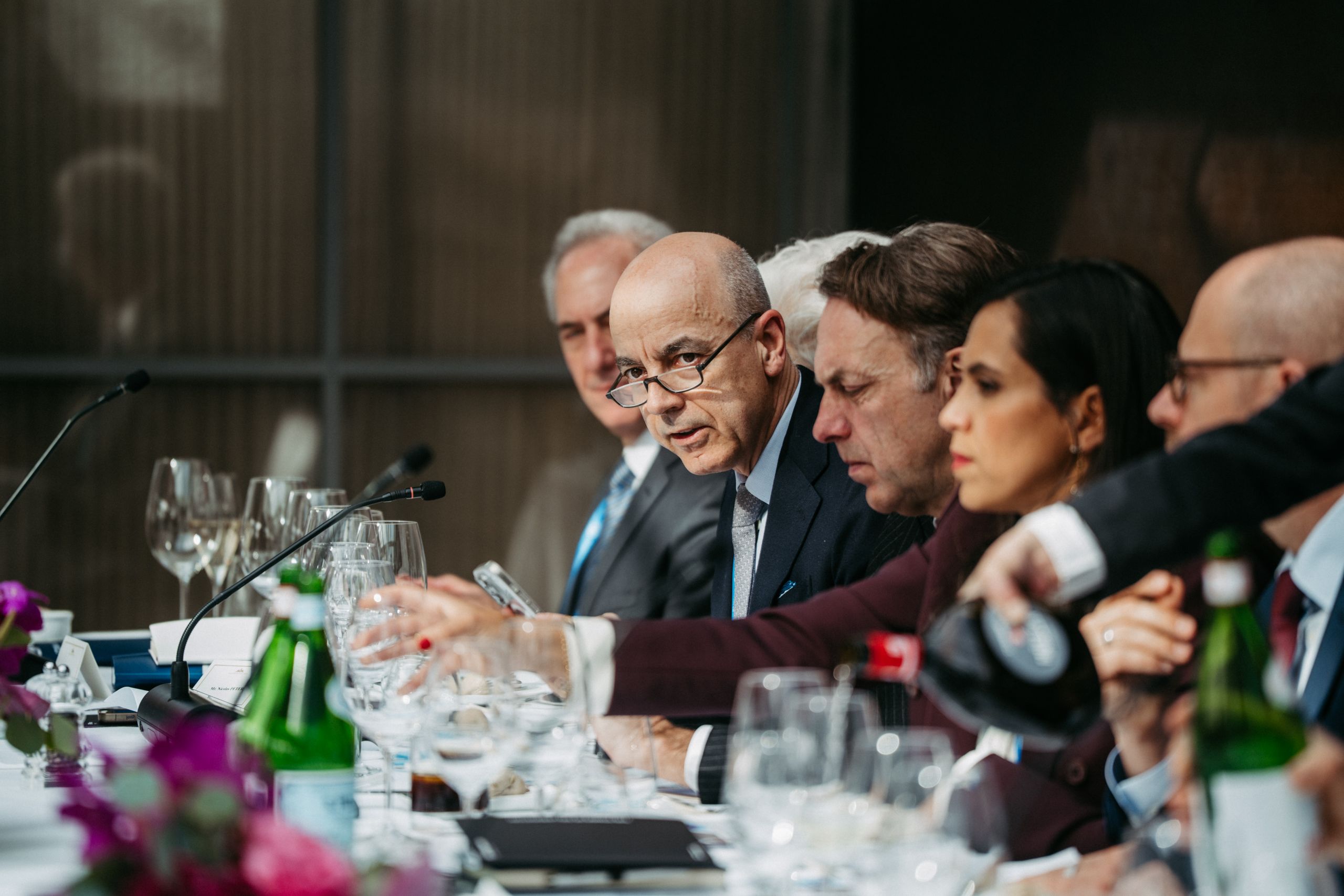 A group of people in formal attire sit at a conference table with documents, microphones, and bottles of water, engaged in discussion.