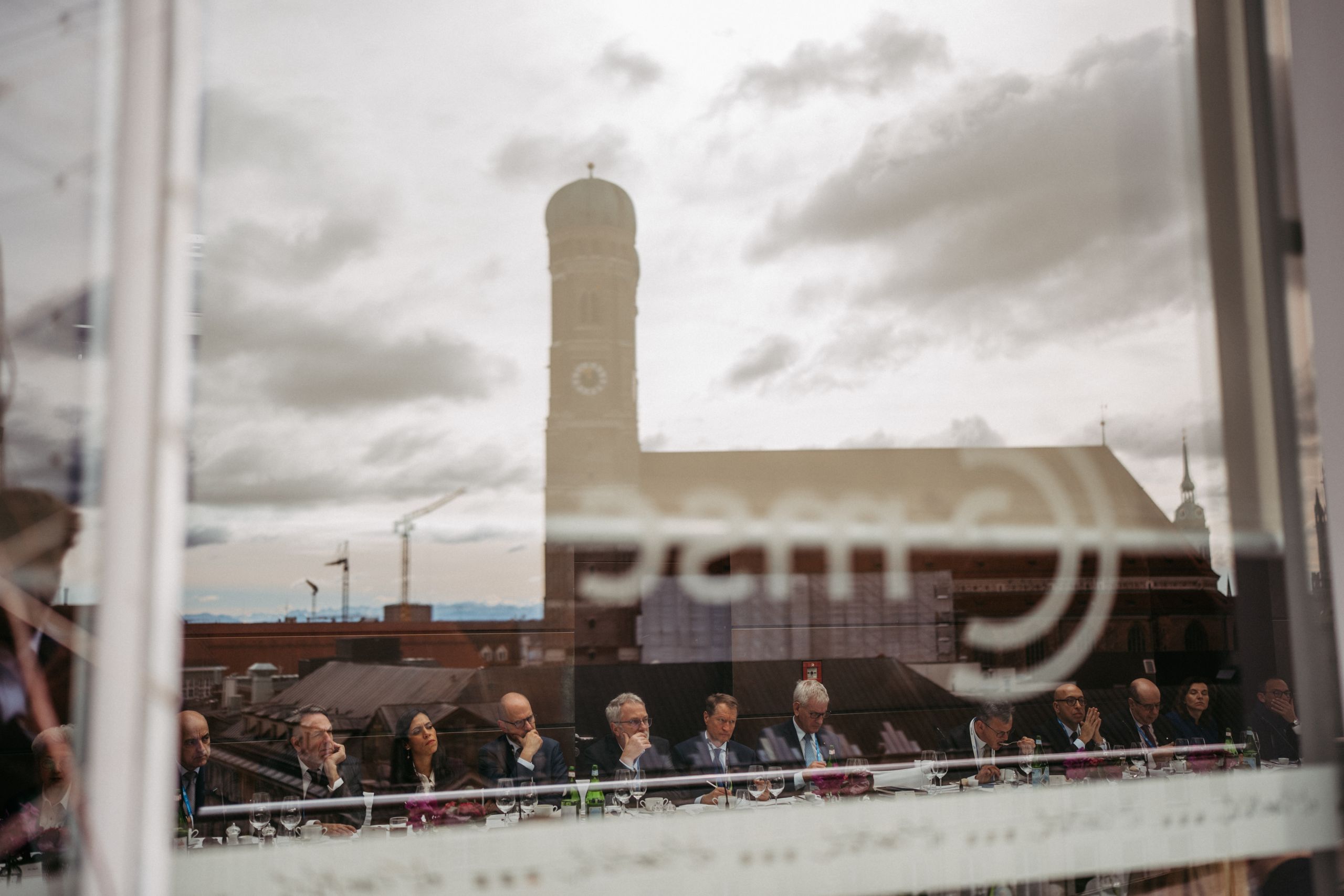 People in a meeting viewed through a window reflecting a cityscape with a church tower and cloudy sky in the background.