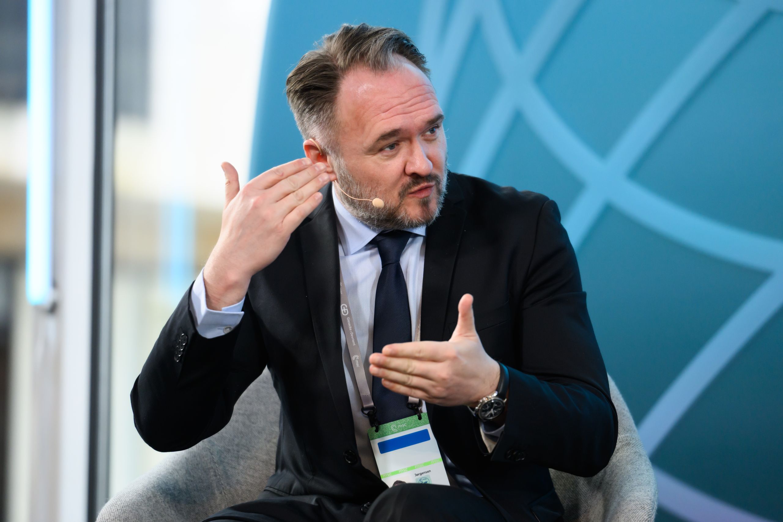 A man in a suit with a headset gestures while speaking in front of a blue globe background, seated and engaged in conversation.