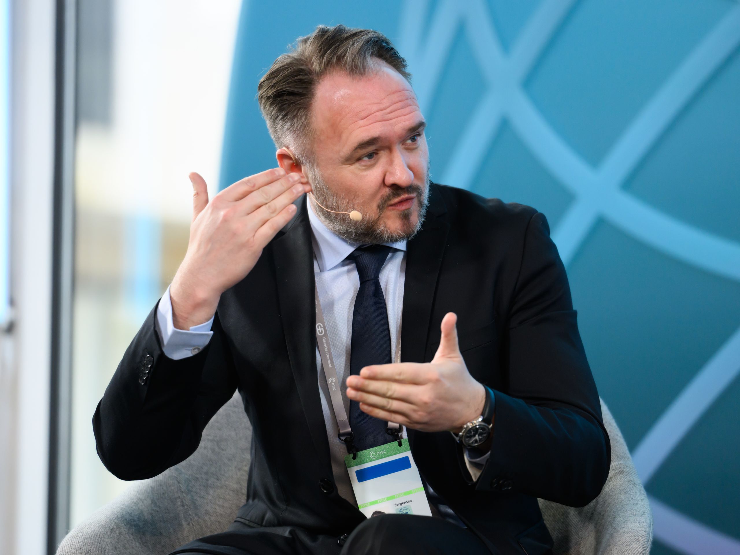 A man in business attire speaks while gesturing with his hands, sitting on a stage with a blue geometric backdrop.