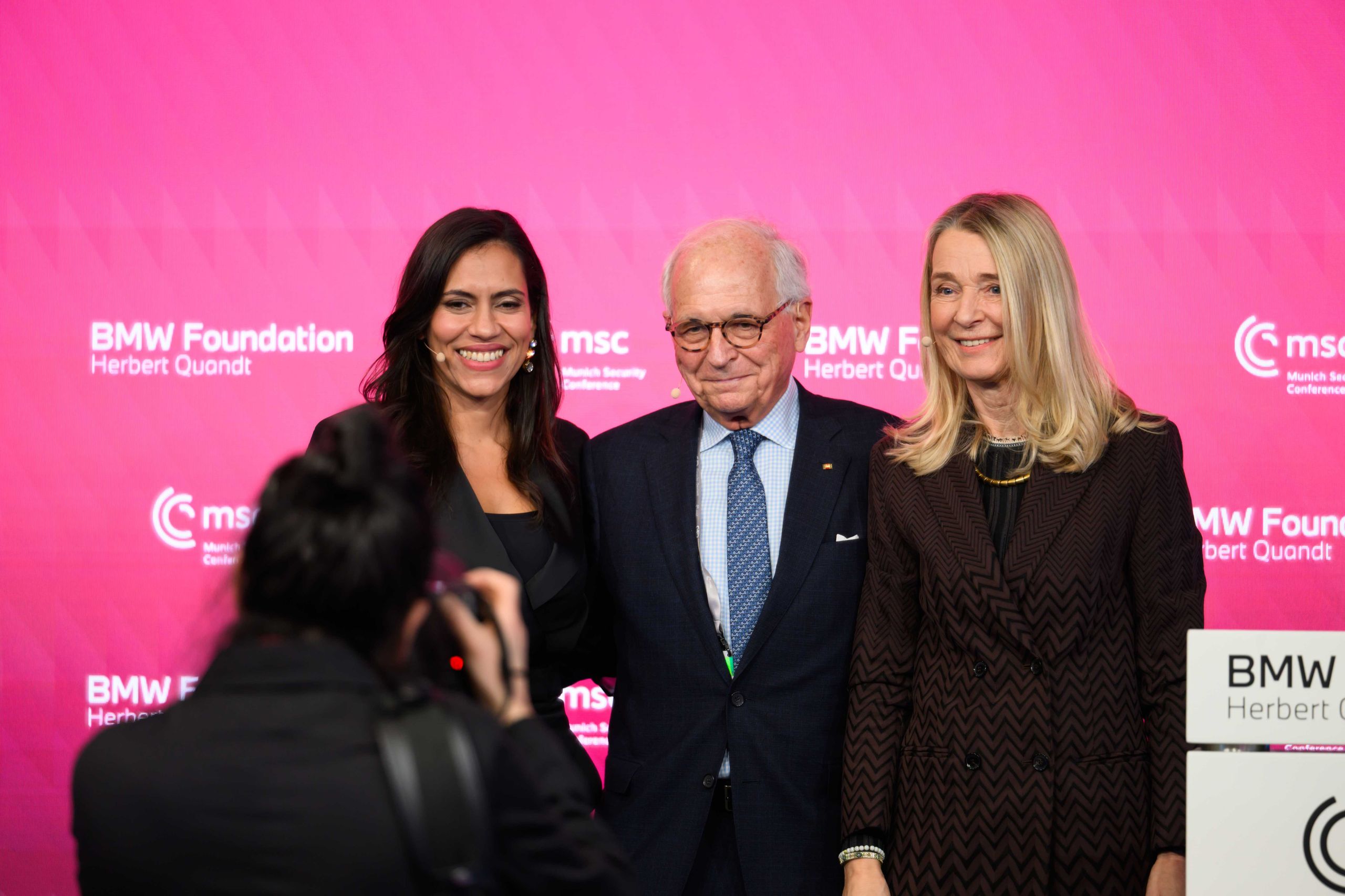 Three people pose for a photo against a pink backdrop with BMW Foundation and MSC logos, as a photographer takes their picture.