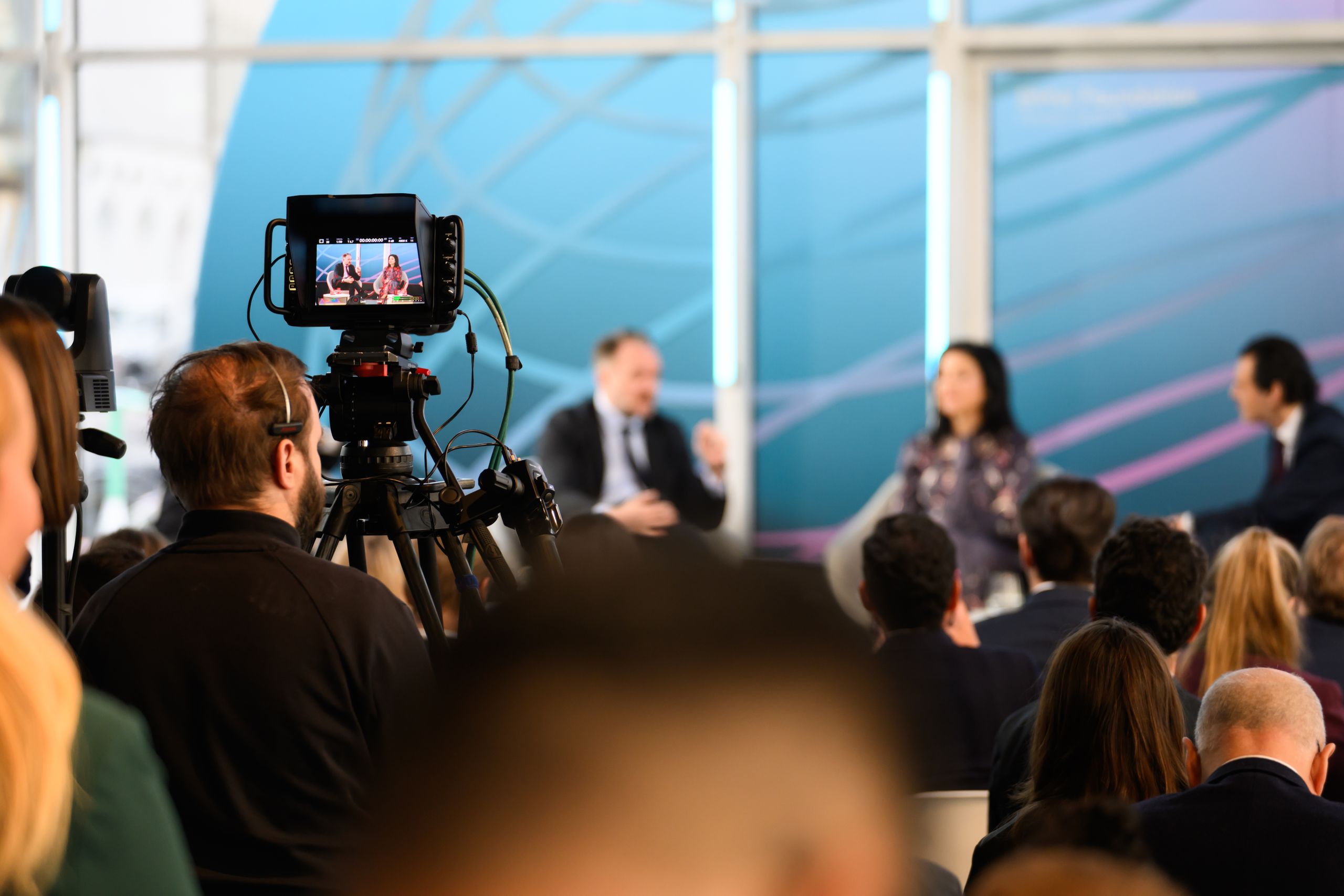 A camera crew films a panel discussion with three speakers on stage, in a brightly lit room with a large audience.