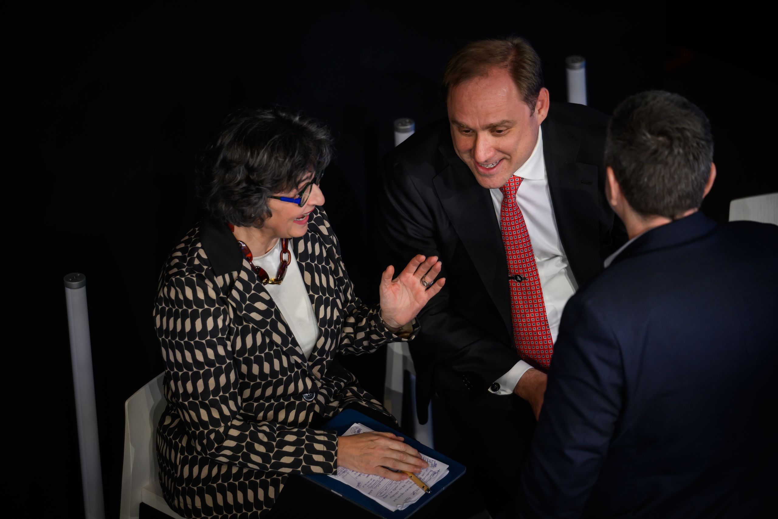 Three people in formal attire are engaged in conversation. A woman gestures while holding papers, and two men listen attentively.