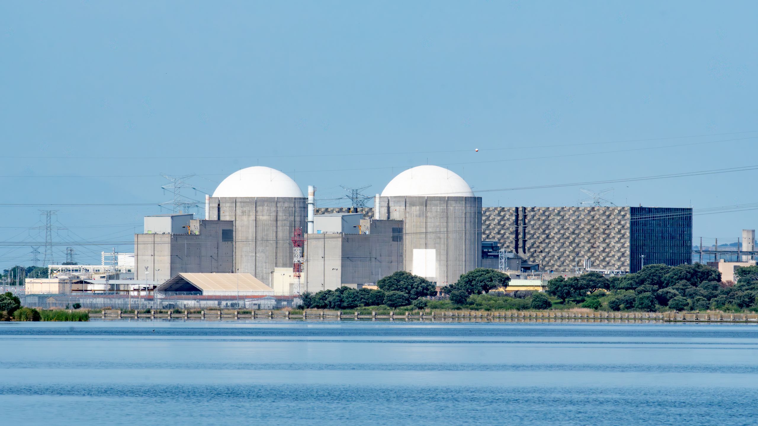 The Almaraz nuclear power plant in Cáceres, Spain, April 2026. Power plant with two large dome-shaped structures by a body of water under a clear blue sky.