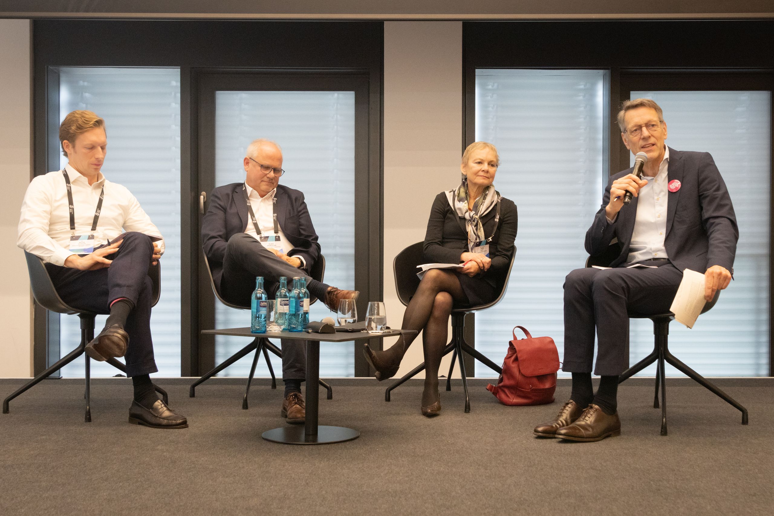 Four people seated on a stage in a panel discussion. One holds a microphone, while others listen attentively. Bottles and glasses on the table.
