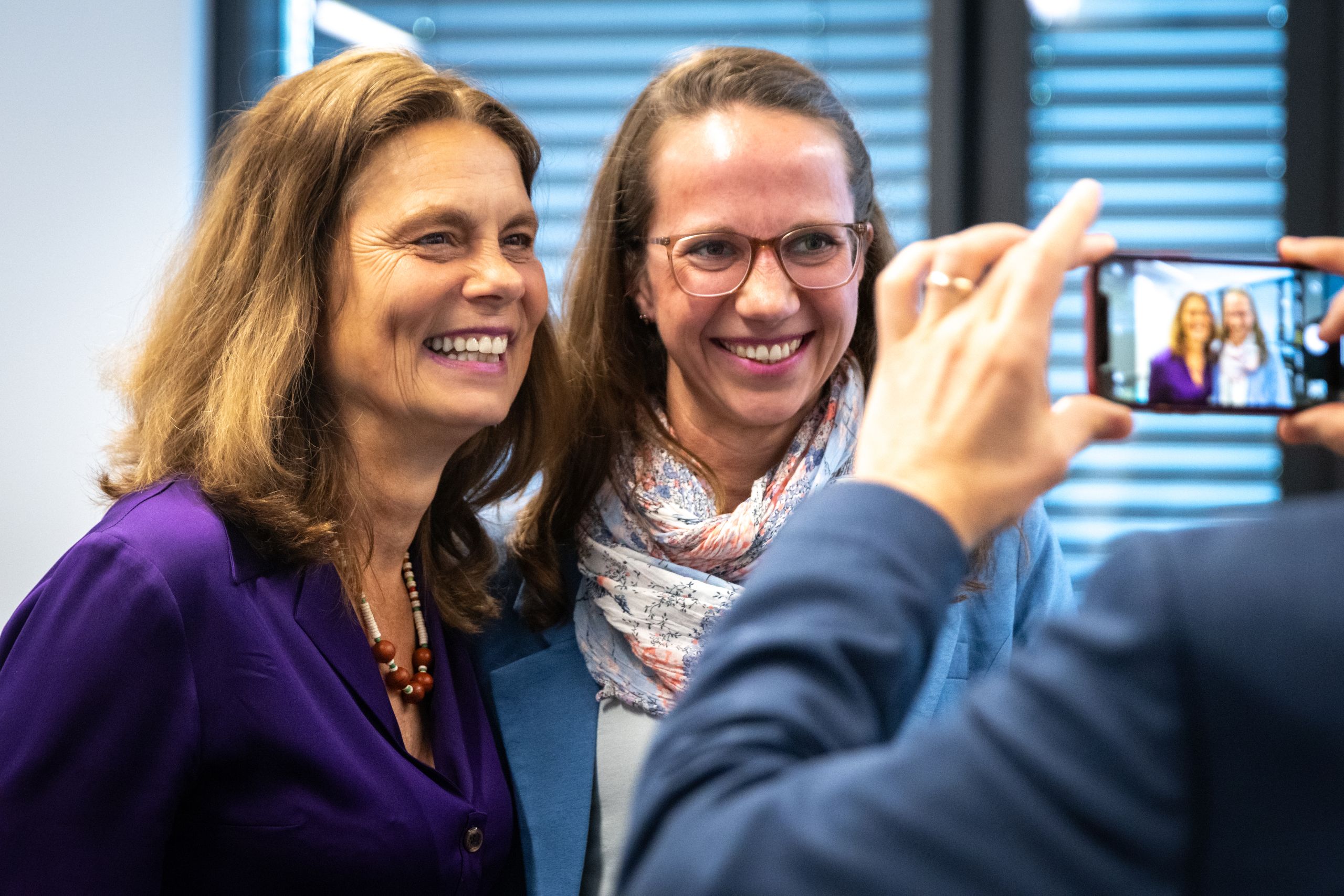 Two women smiling for a photo, one in a purple top and the other in glasses and a scarf. A person in blue takes their picture with a phone.