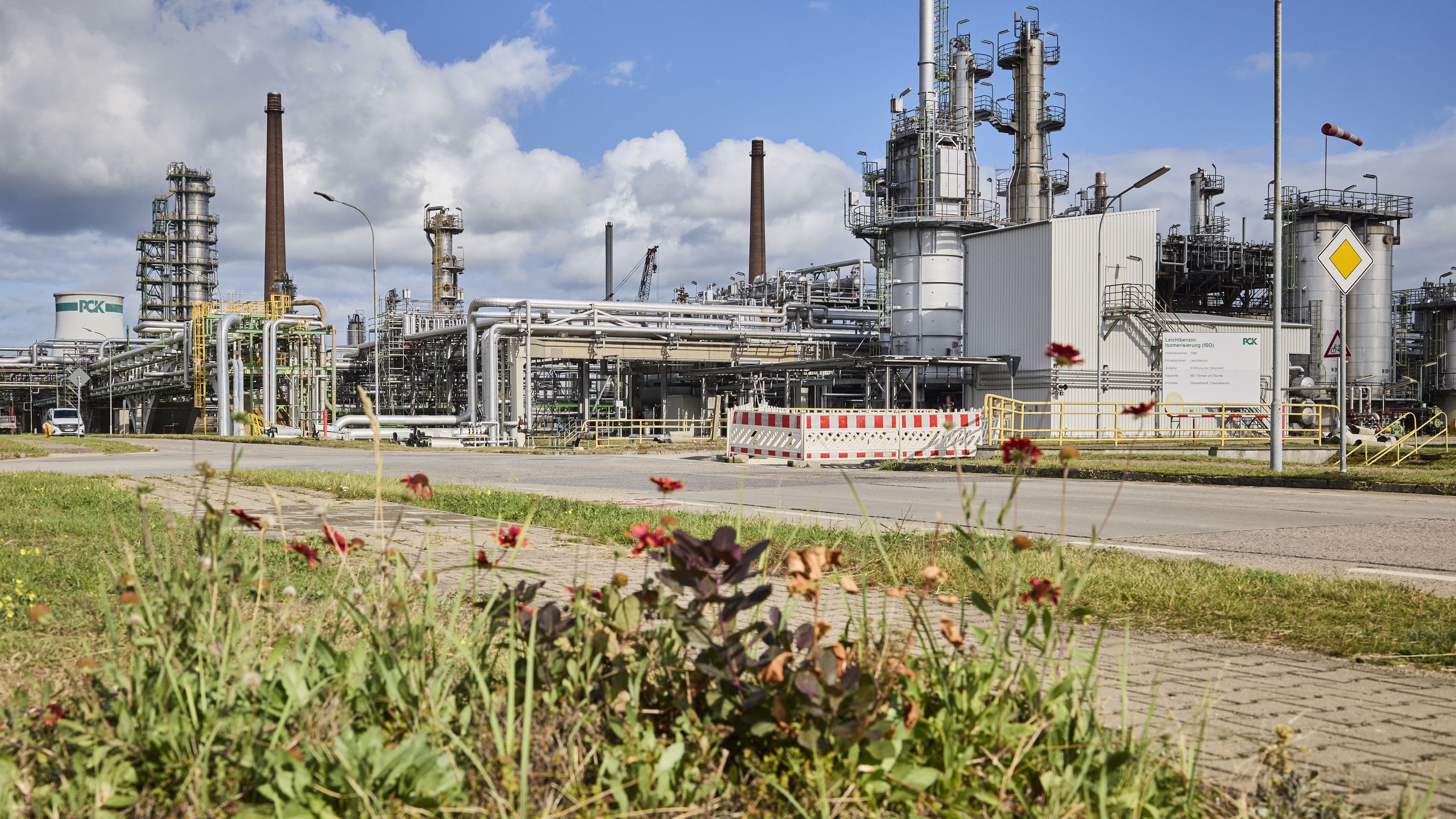 The PCK refinery in Schwedt, Germany, faces the challenge of transitioning from fossil fuels to cleaner energy sources. Industrial facility with pipes and towers under a blue sky, with flowering plants in the foreground.