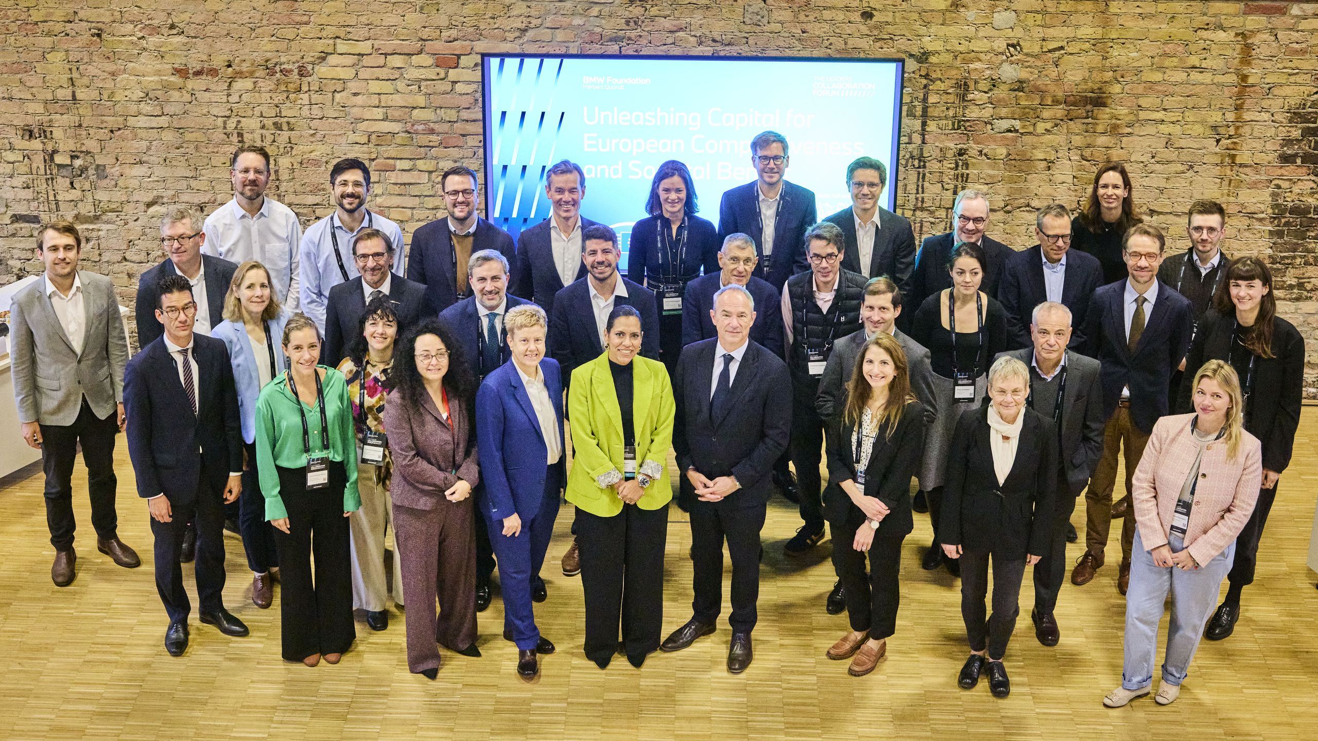 Participants at the roundtable "Unleashing Capital for European Competitiveness and Societal Benefit". A group of 30 people, men and women, standing indoors in front of a brick wall and a large screen, smiling and posing for a photo.