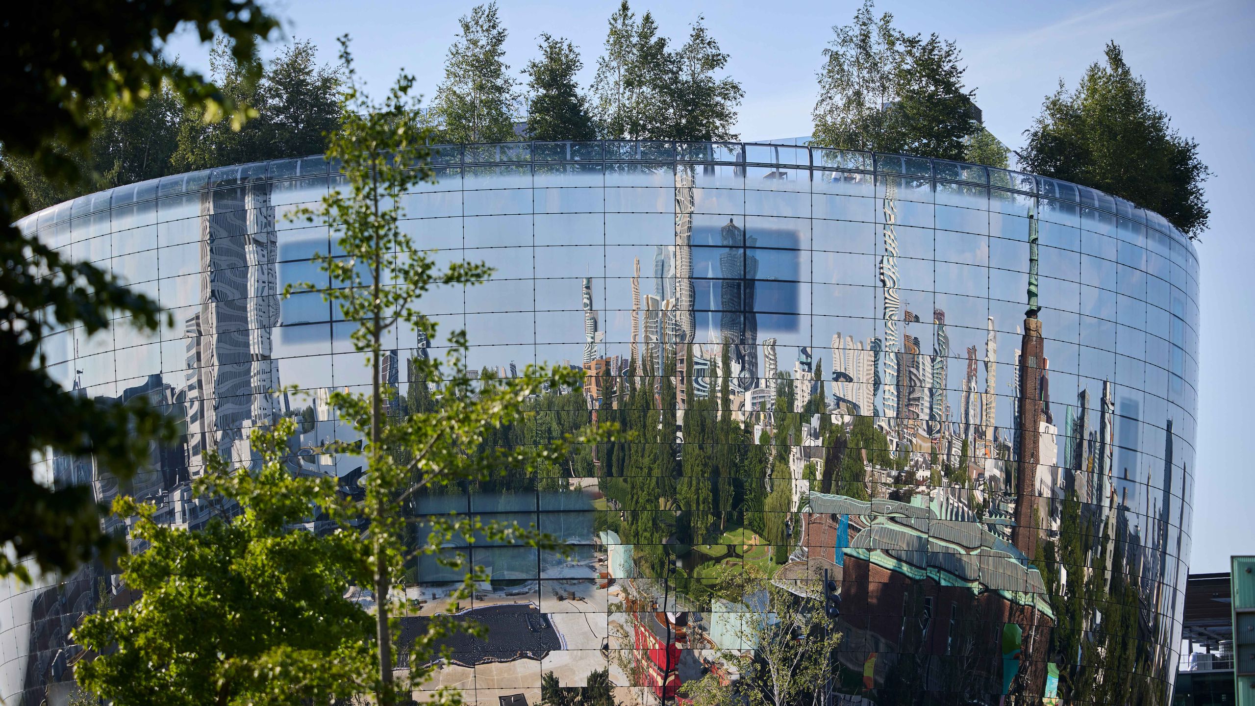 title A circular glass building with trees on the roof reflects a distorted cityscape, surrounded by greenery under a clear blue sky.