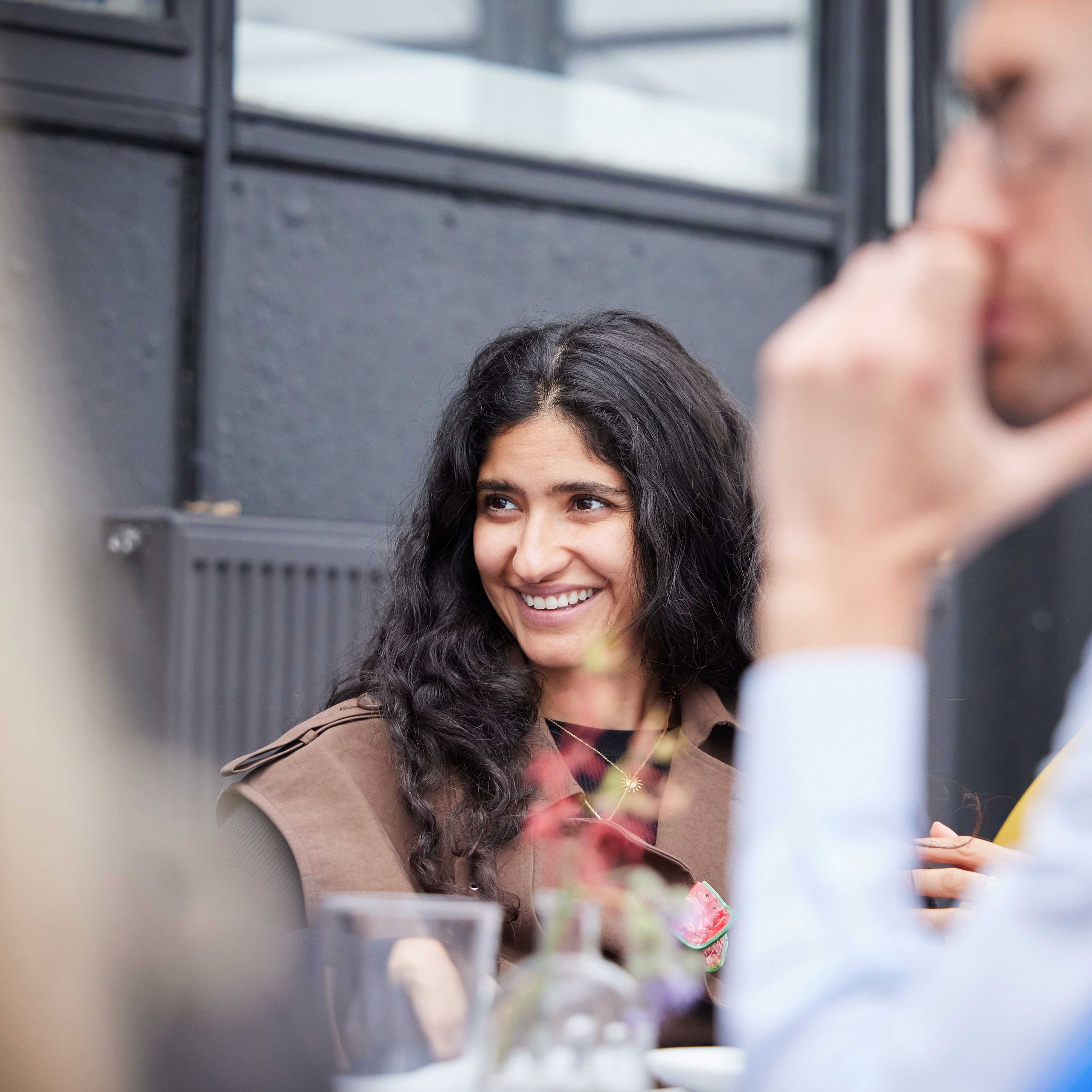 A woman with long dark hair smiles while sitting at a table during a social gathering. Other people are blurred in the foreground.