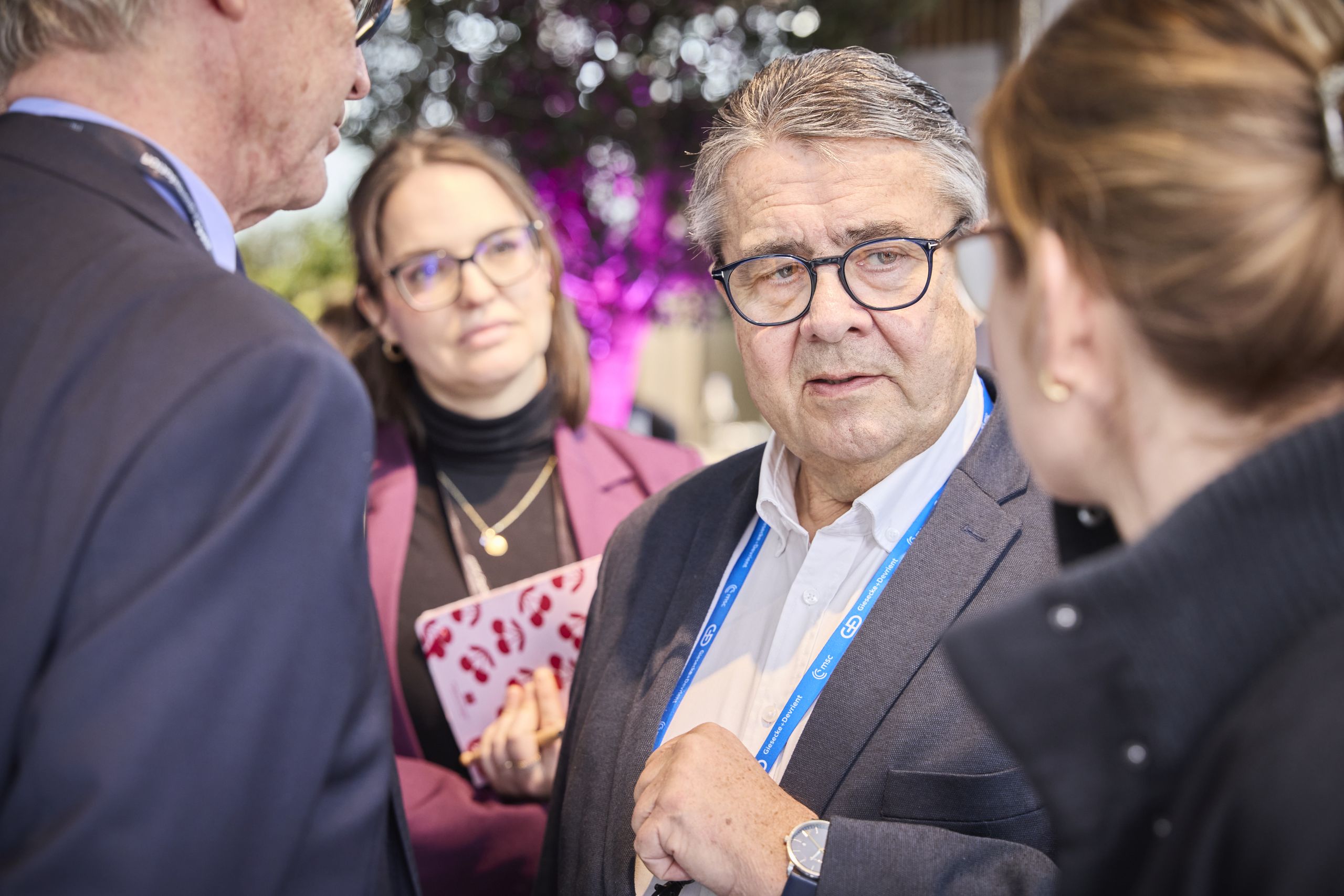 A group of people in conversation indoors; a man with glasses and a blue lanyard speaks while others listen attentively.