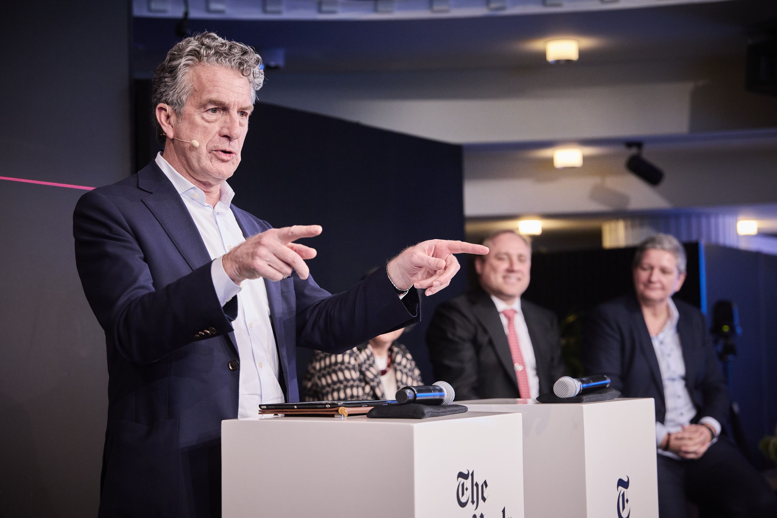 Man speaking at a podium during a panel discussion, with three people seated behind him and microphones on the podium.