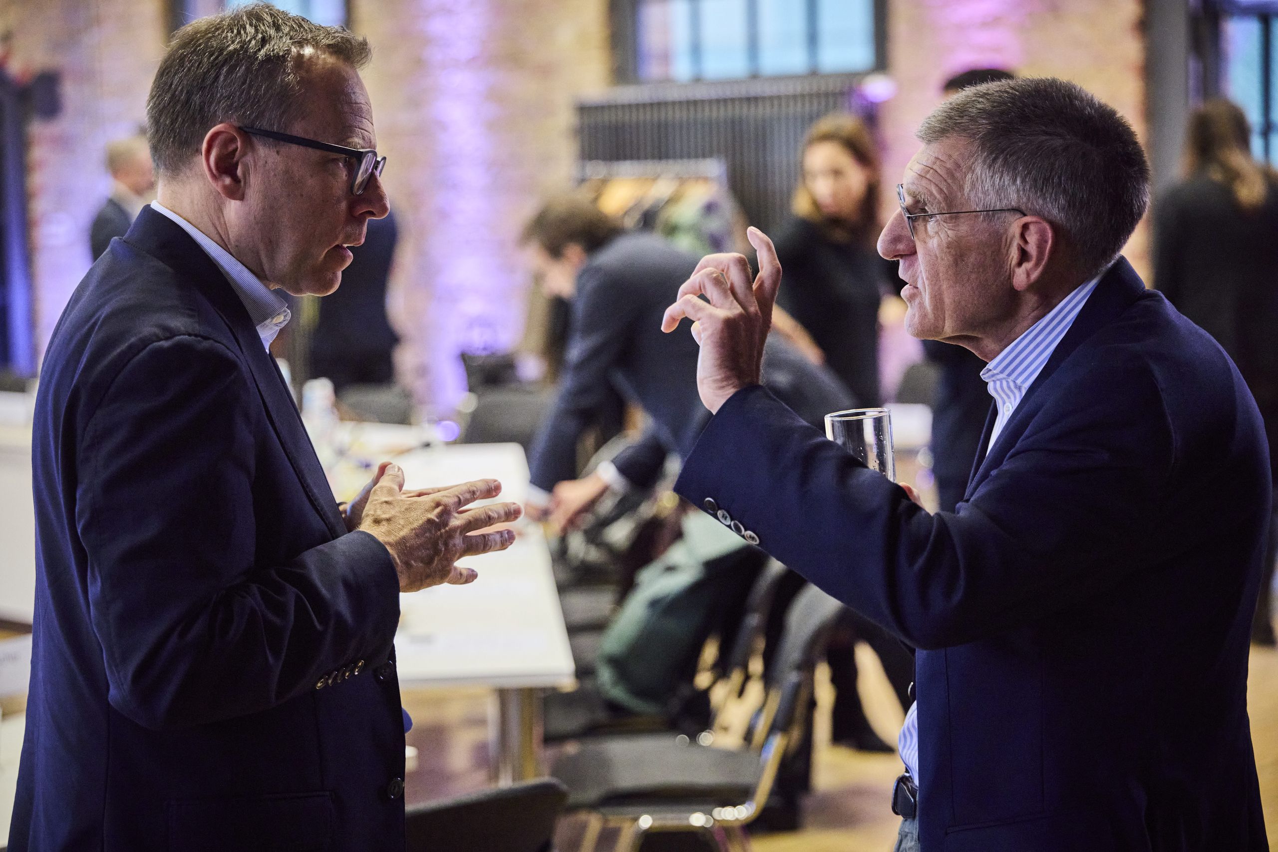 Participants engaged in vivid discussions. Two men engaged in conversation at an indoor event, with others in the background and tables with chairs nearby.