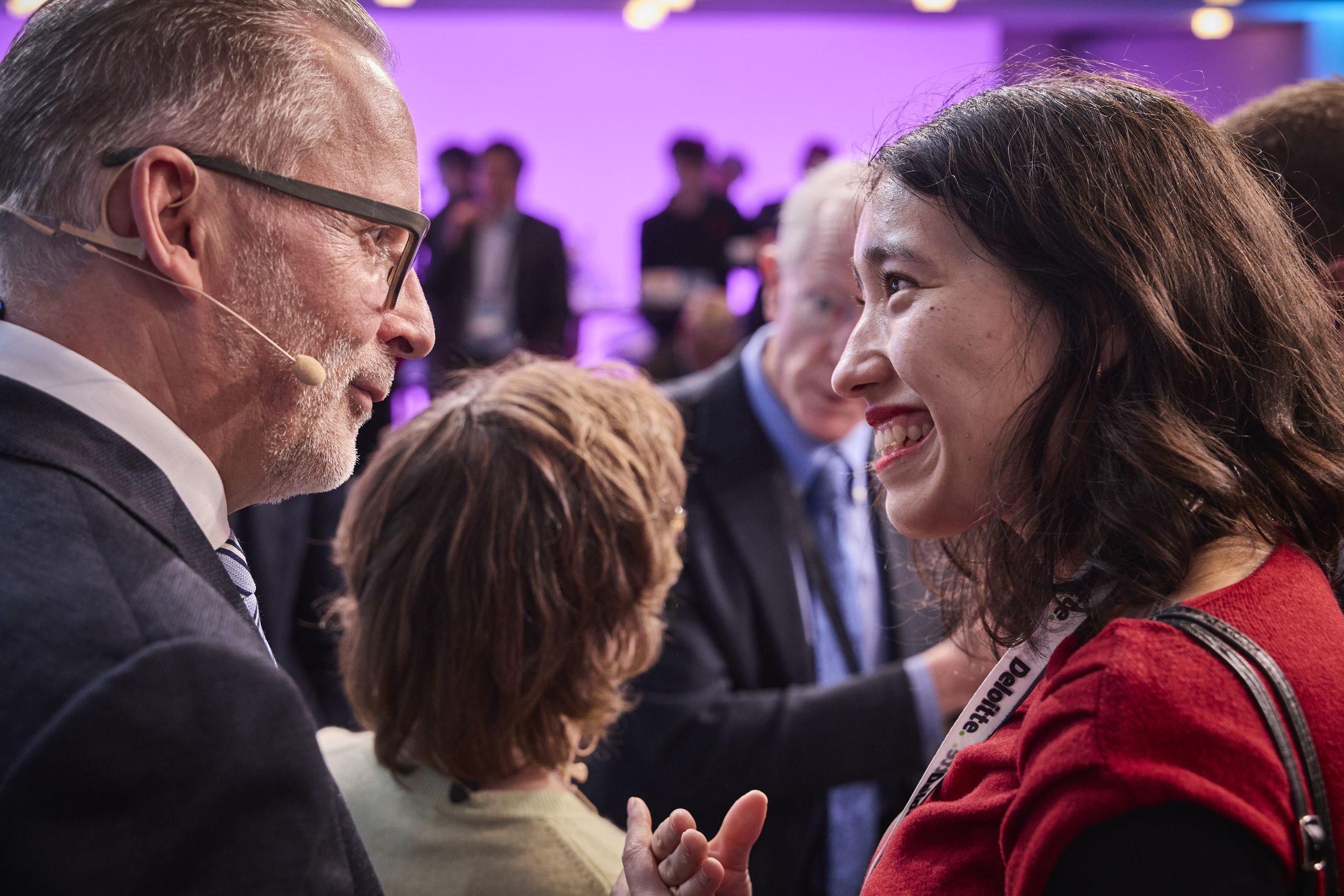 Two people smiling and talking at a conference, wearing business attire. A crowd is visible in the blurred background under purple lighting.