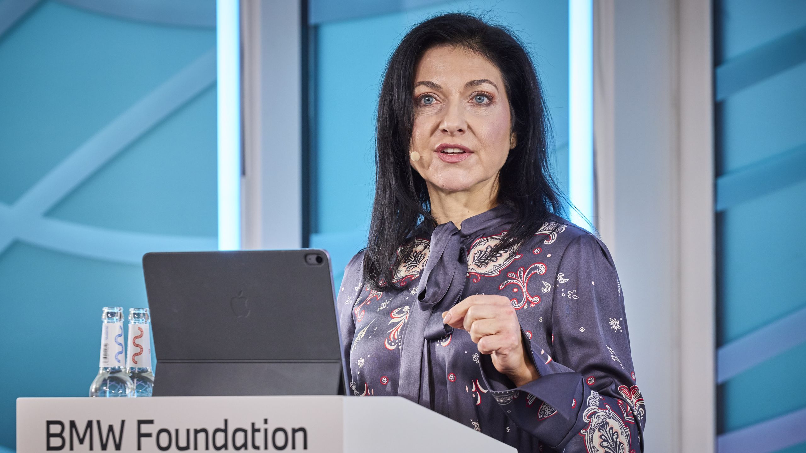 A woman speaks at a podium during a BMW Foundation event, standing in front of a turquoise backdrop.