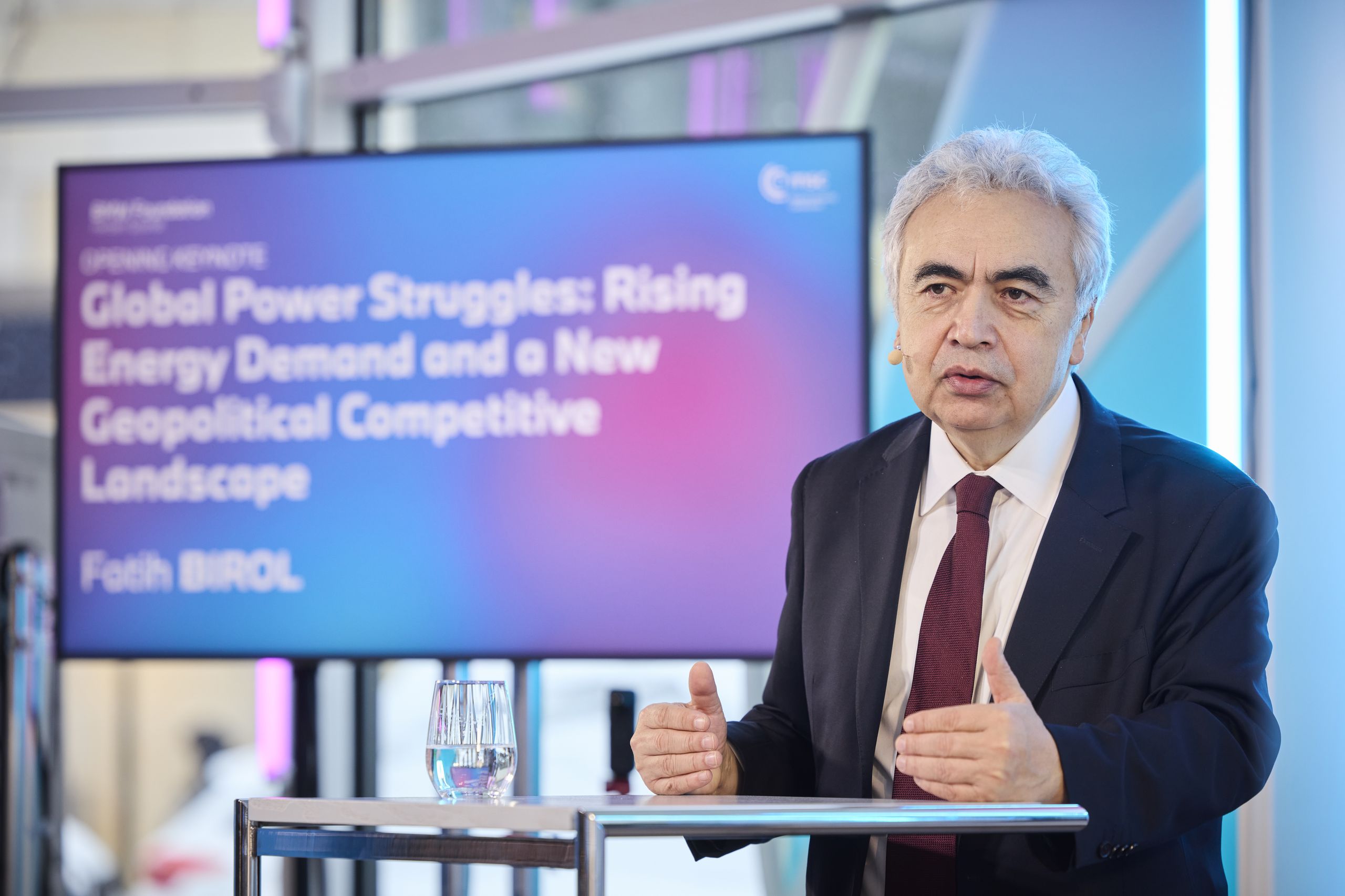 Man in a suit speaks at an event with a screen behind him displaying a presentation title about global energy and geopolitical issues.