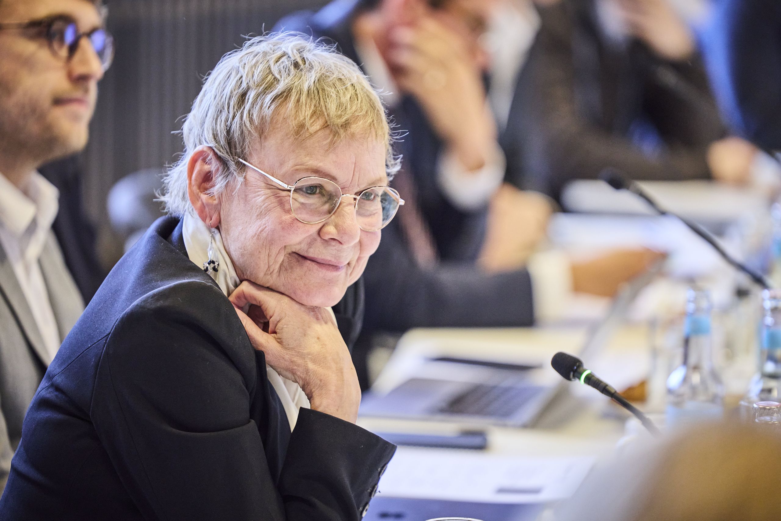 Sabine Kunst, Chair of the Board of the Hamburg-based Joachim Herz Stiftung. An elderly person with glasses smiles while sitting at a conference table, surrounded by others, with a laptop and microphone in view.